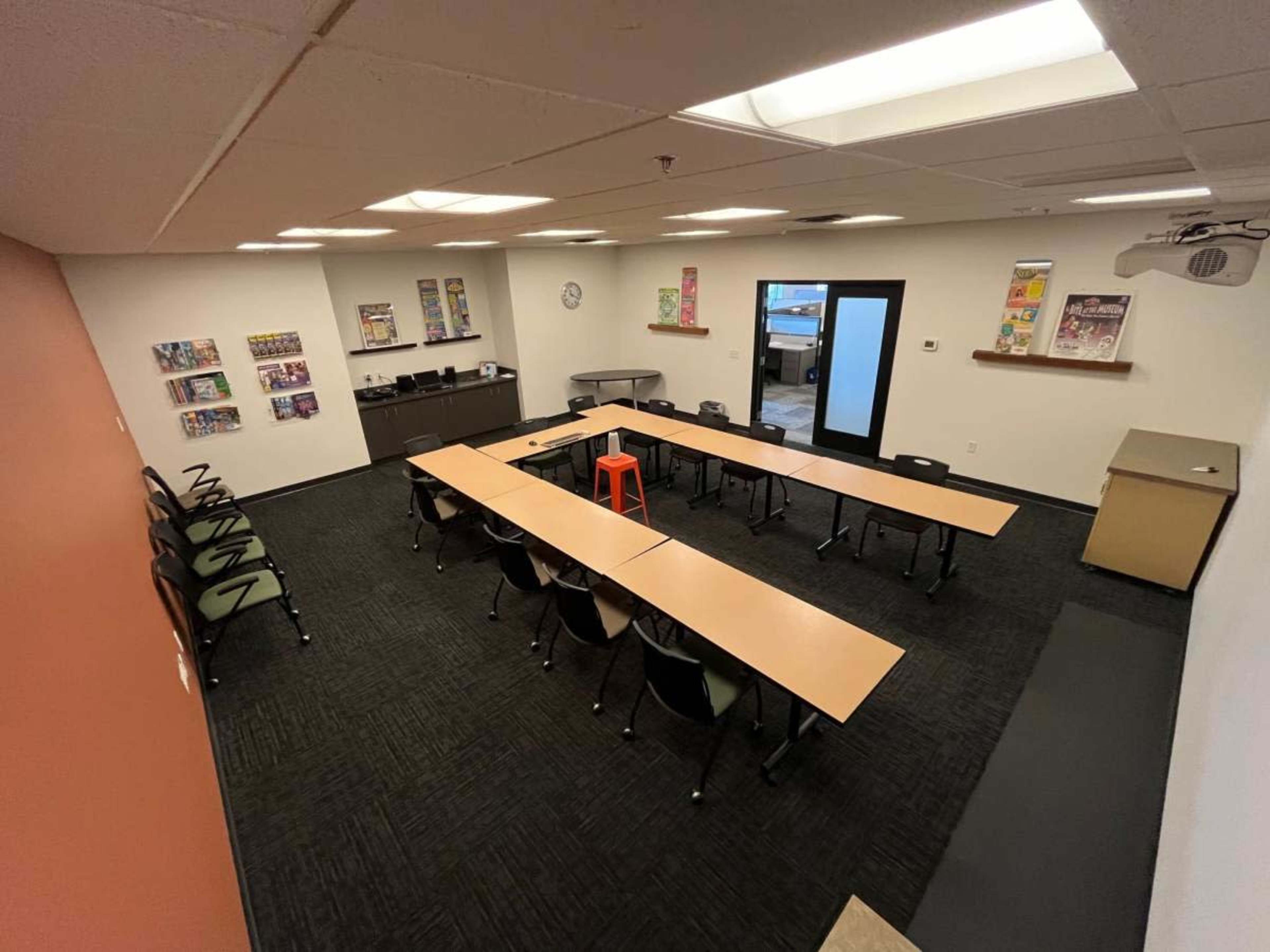 A meeting room with several tables arranged in a U-shape, black chairs, and a single orange stool in the center.