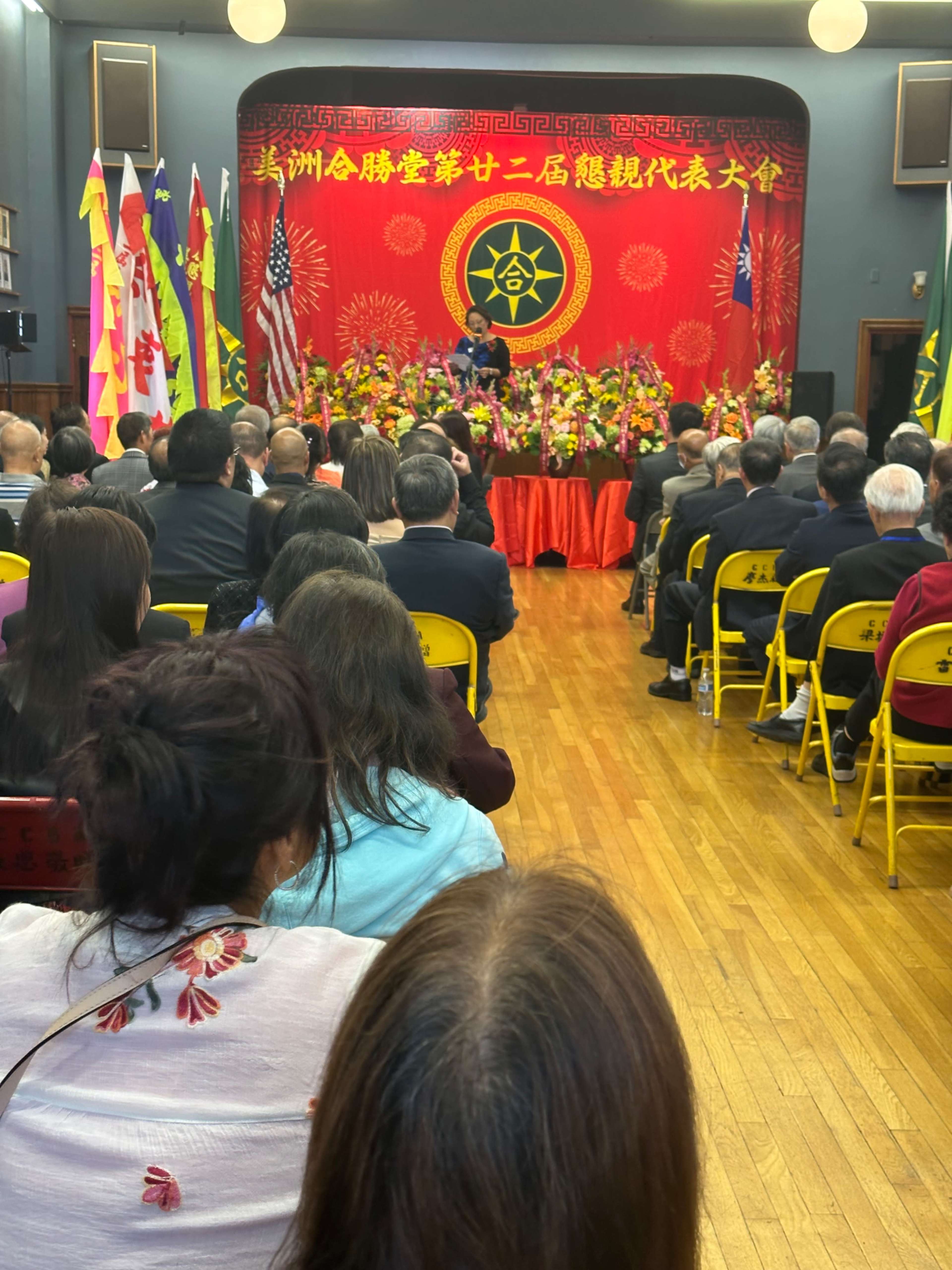 A speaker addresses an audience at a formal event decorated with flags and flowers in a venue featuring a red backdrop.