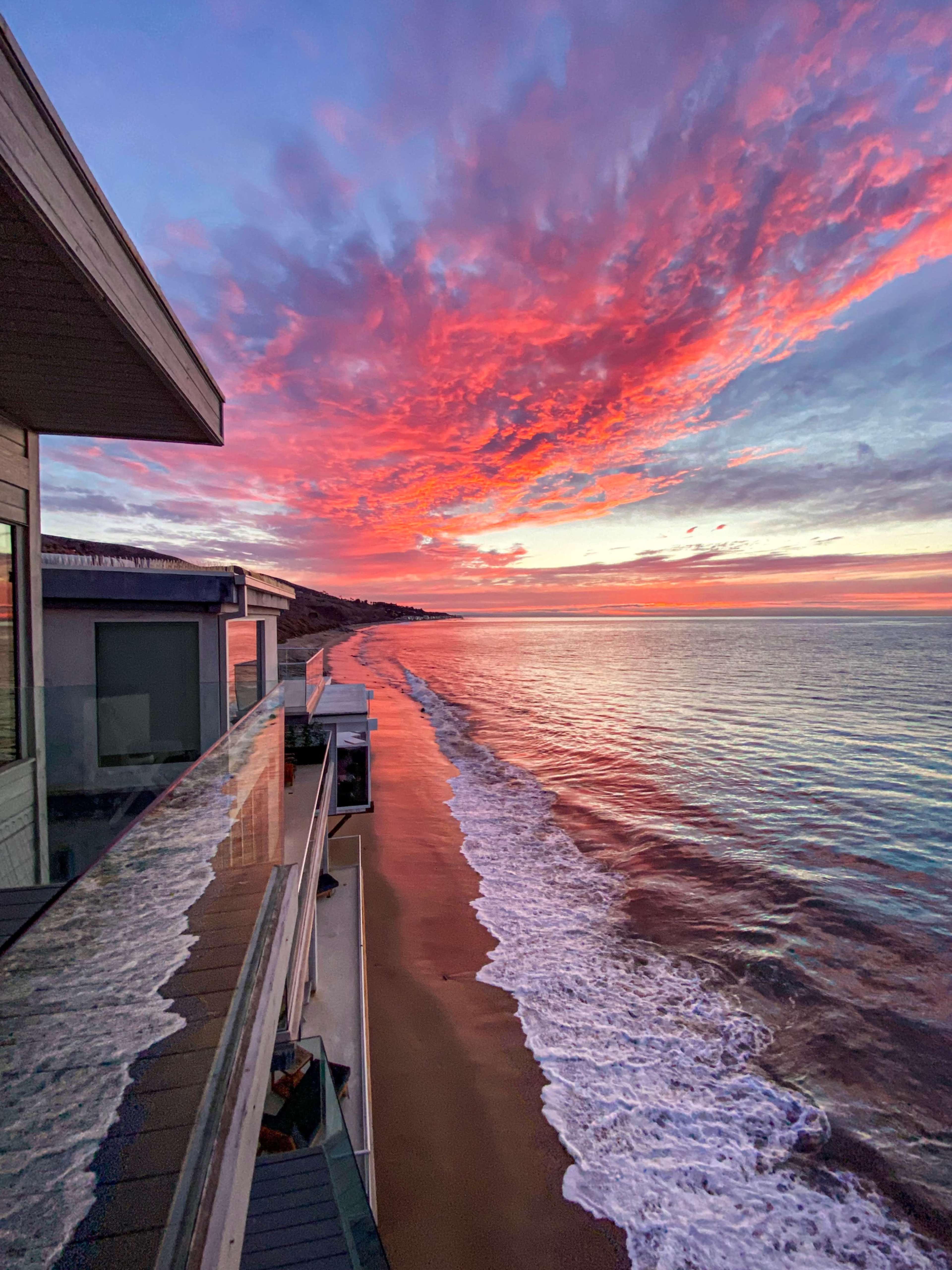 A vibrant sunset casts colorful reflections over a calm ocean along a sandy beach, with a modern building on the left.