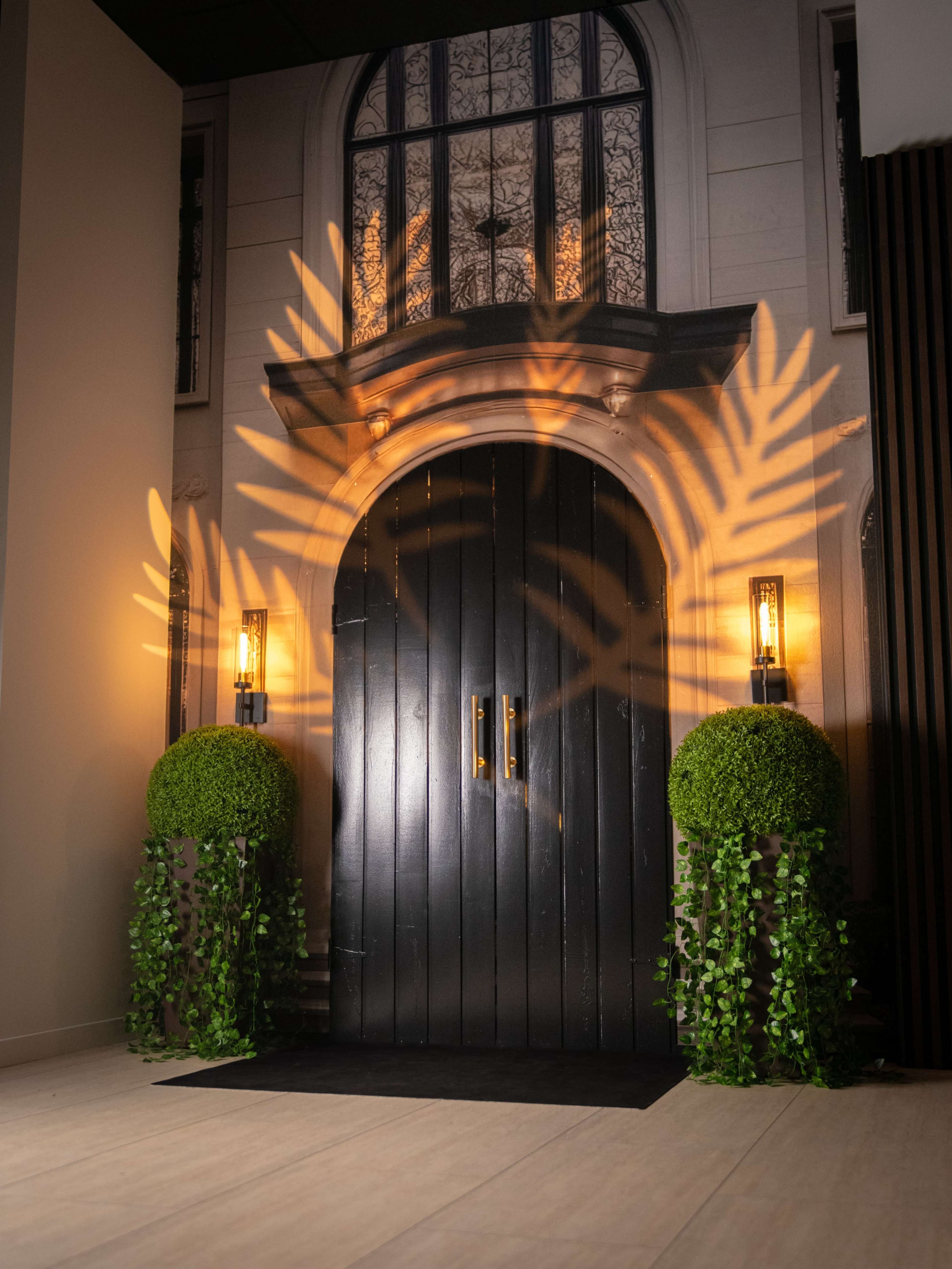 The image shows a decorative entrance with a large black door framed by two green topiary plants and warm, wall-mounted lights casting leaf-shaped shadows.