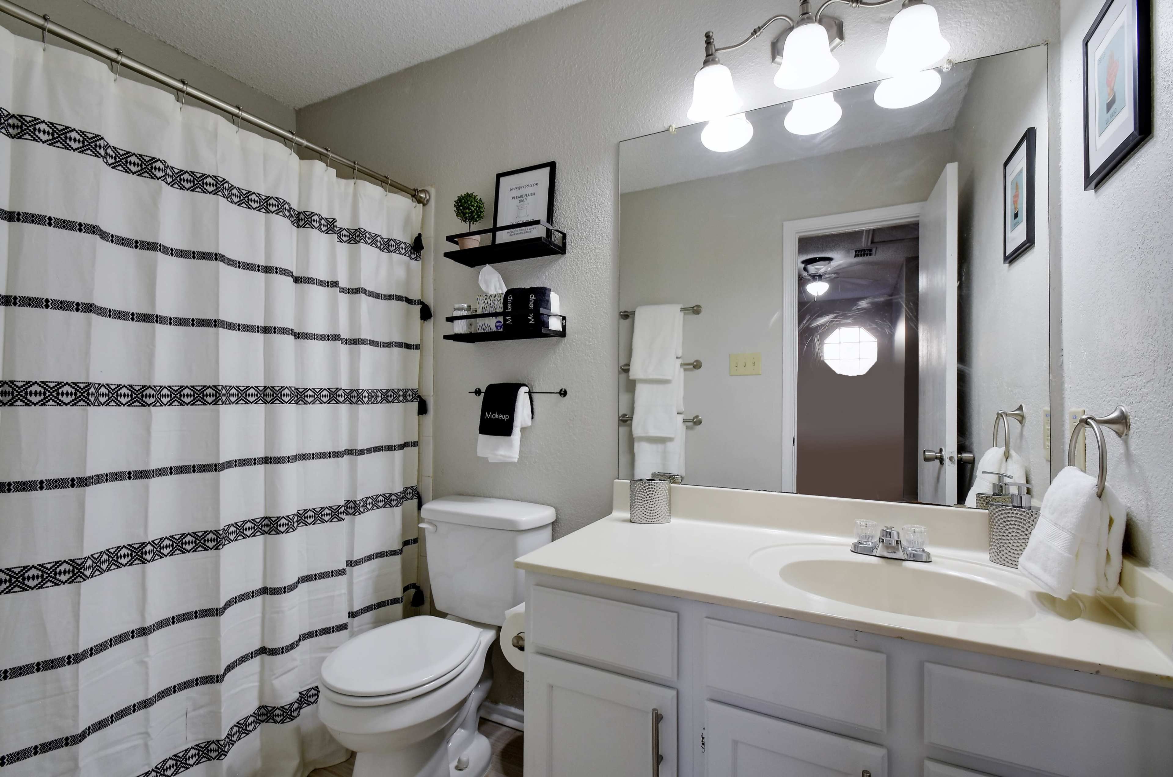 The image shows a bathroom with a shower curtain, a toilet, a sink with a countertop, and shelves displaying toiletries.