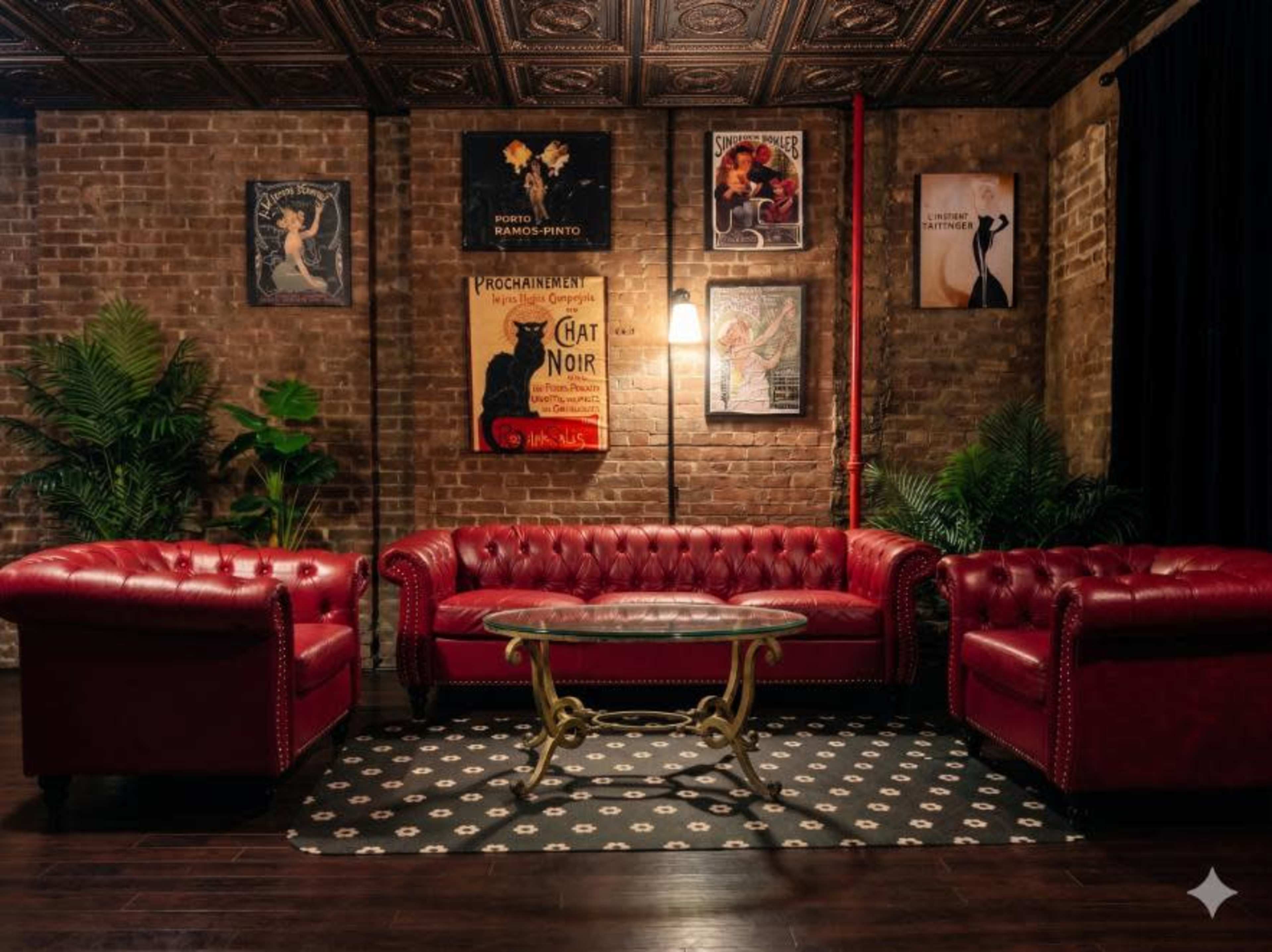 The image features a vintage-style lounge area with a red leather chesterfield sofa and matching armchairs, a glass-top coffee table, and decorative posters on a brick wall.