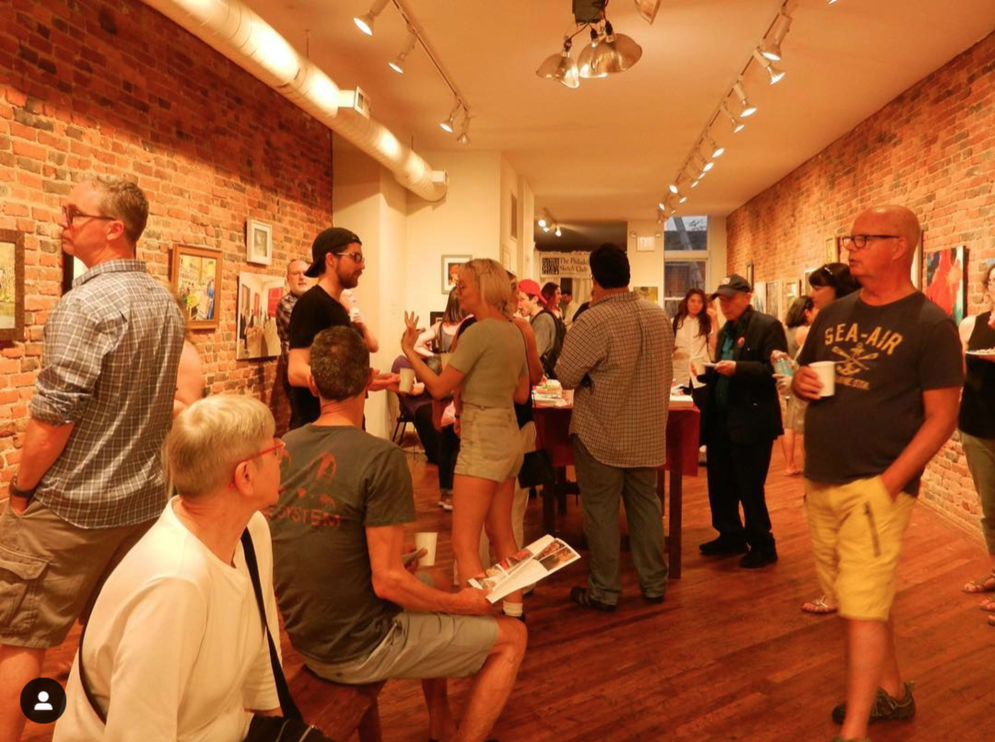 A group of people is gathered in an art gallery featuring exposed brick walls and various artworks on display.