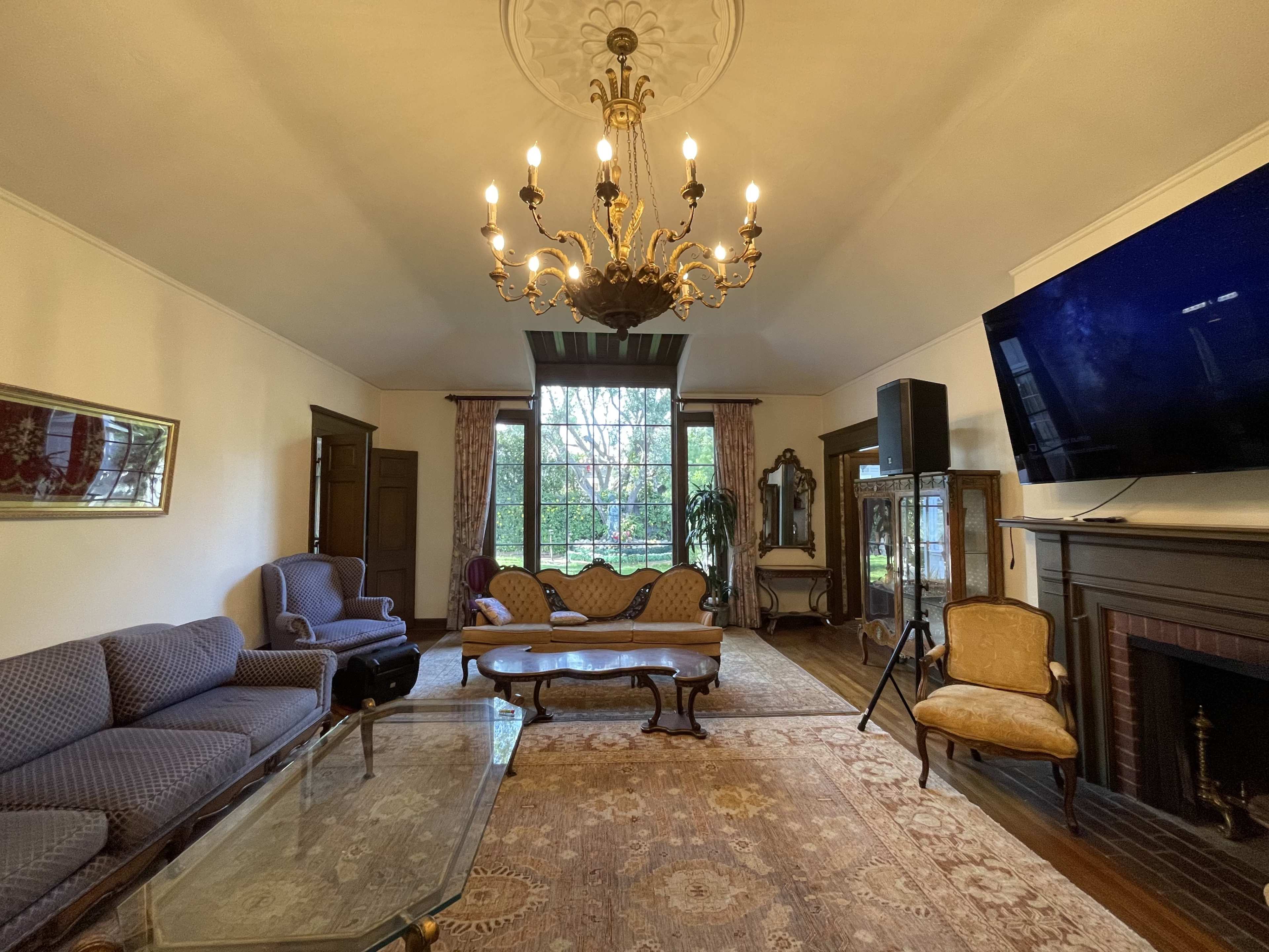 A spacious living room with a chandelier, a large window, and various pieces of vintage furniture, including a sofa, armchairs, and a coffee table.