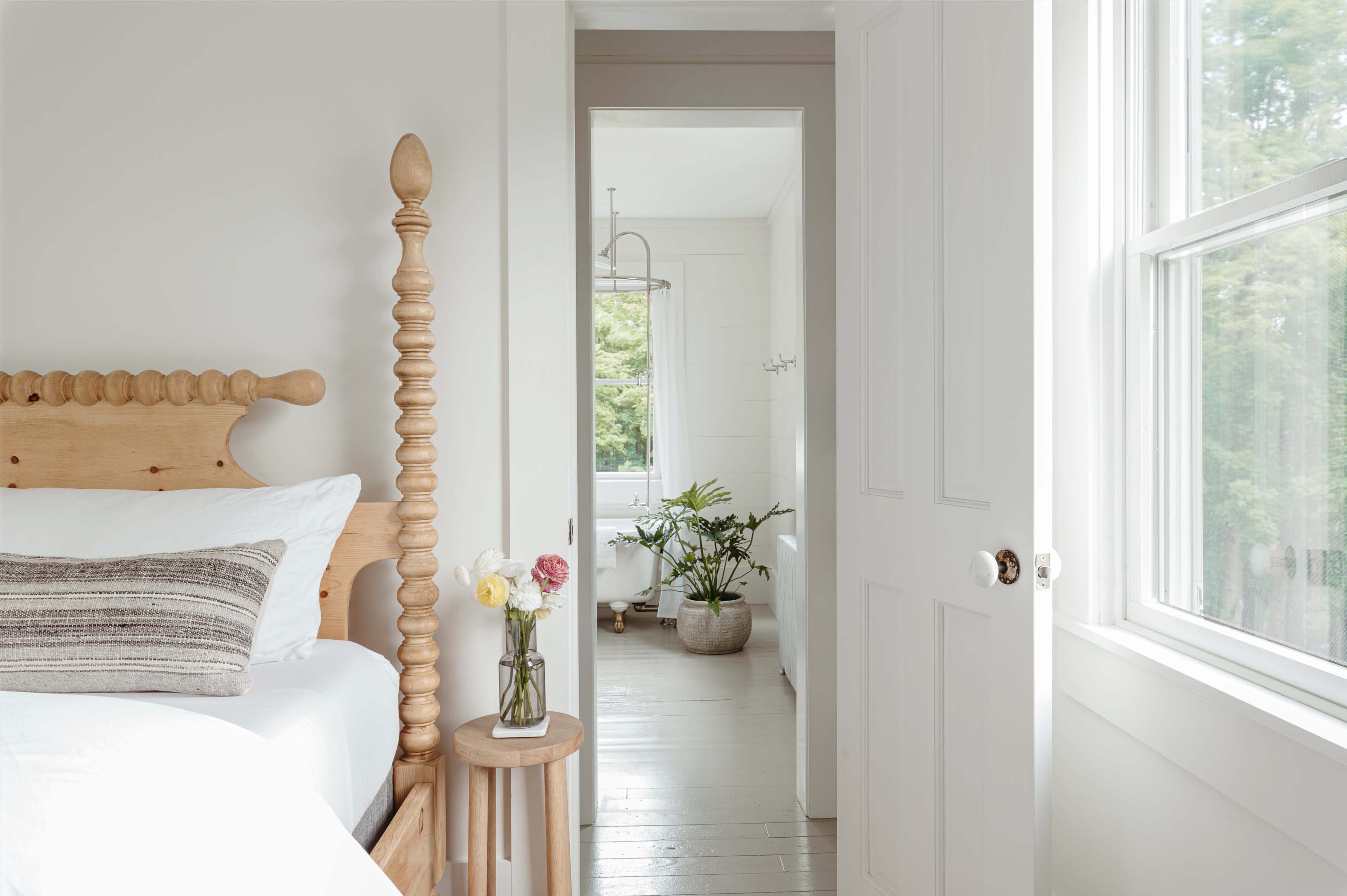 A light-filled bedroom features a wooden bed with a decorative headboard, a bedside table with a vase of flowers, and an open door leading to a bathroom with plants visible.