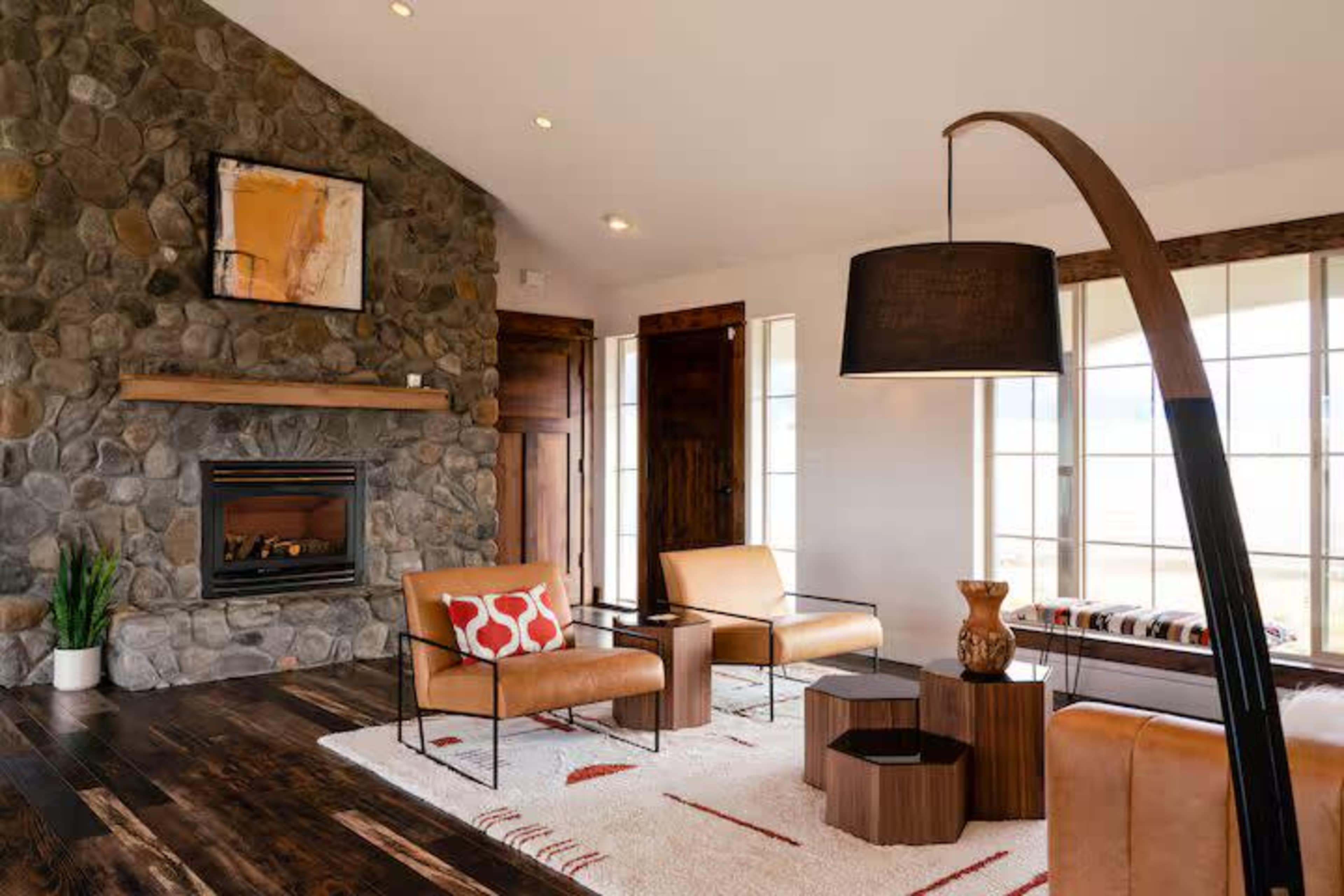 The image shows a modern living room featuring a stone fireplace, two orange armchairs, a wooden coffee table, and large windows allowing natural light.