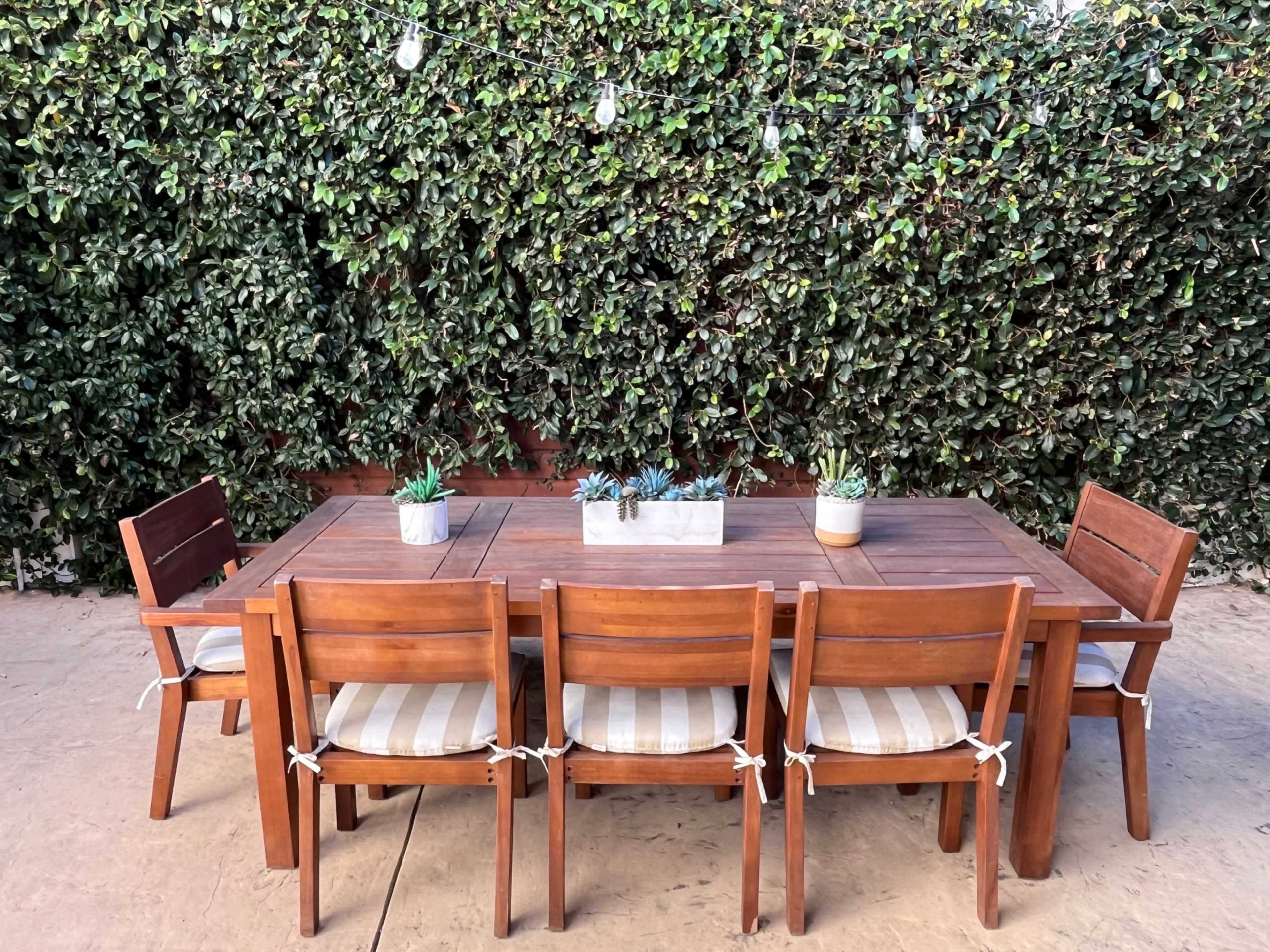 A wooden outdoor dining table is surrounded by six chairs against a backdrop of dense greenery.