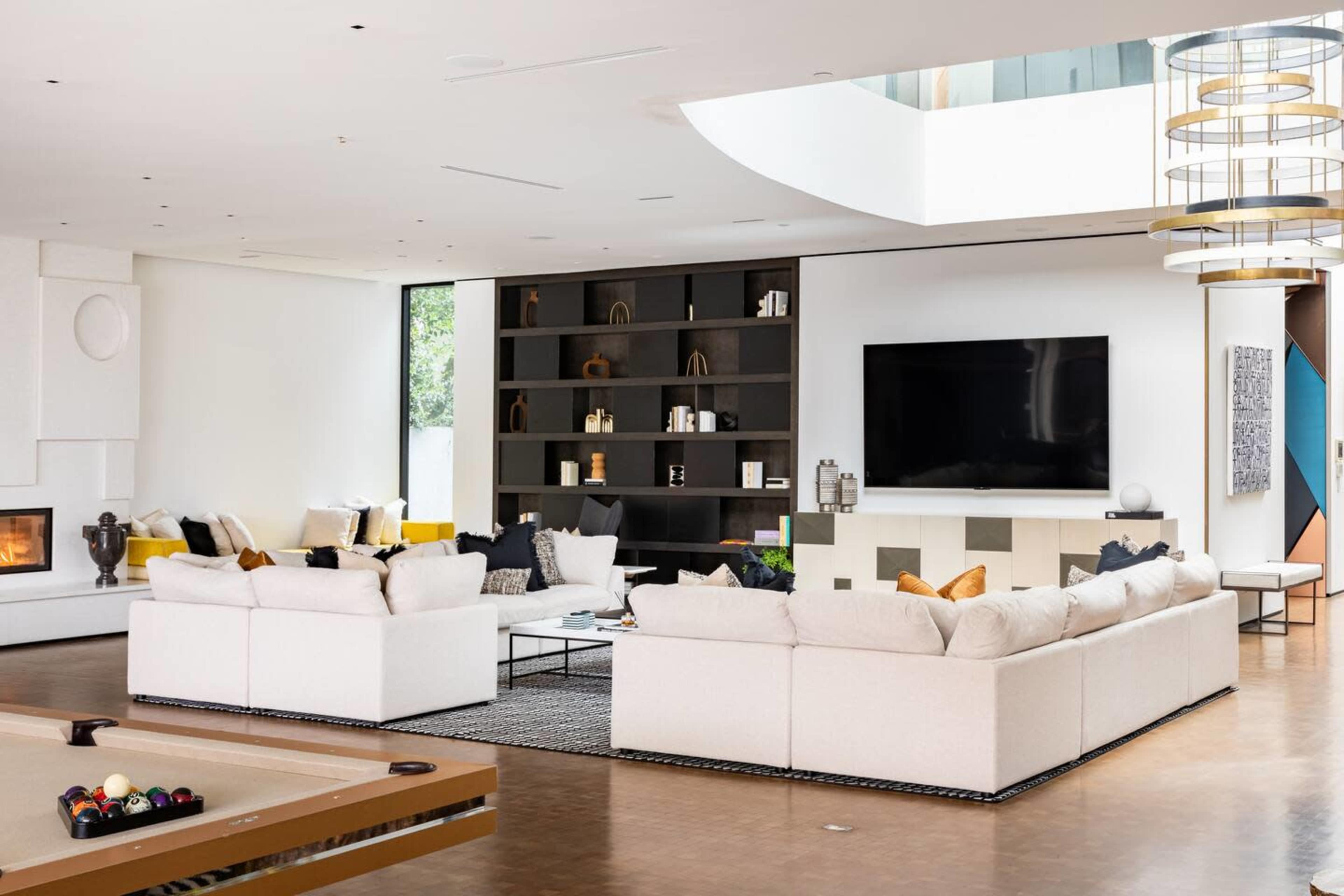 A spacious living room features white sofas arranged around a coffee table, with a large TV mounted on a black and white entertainment unit, and a modern chandelier overhead.