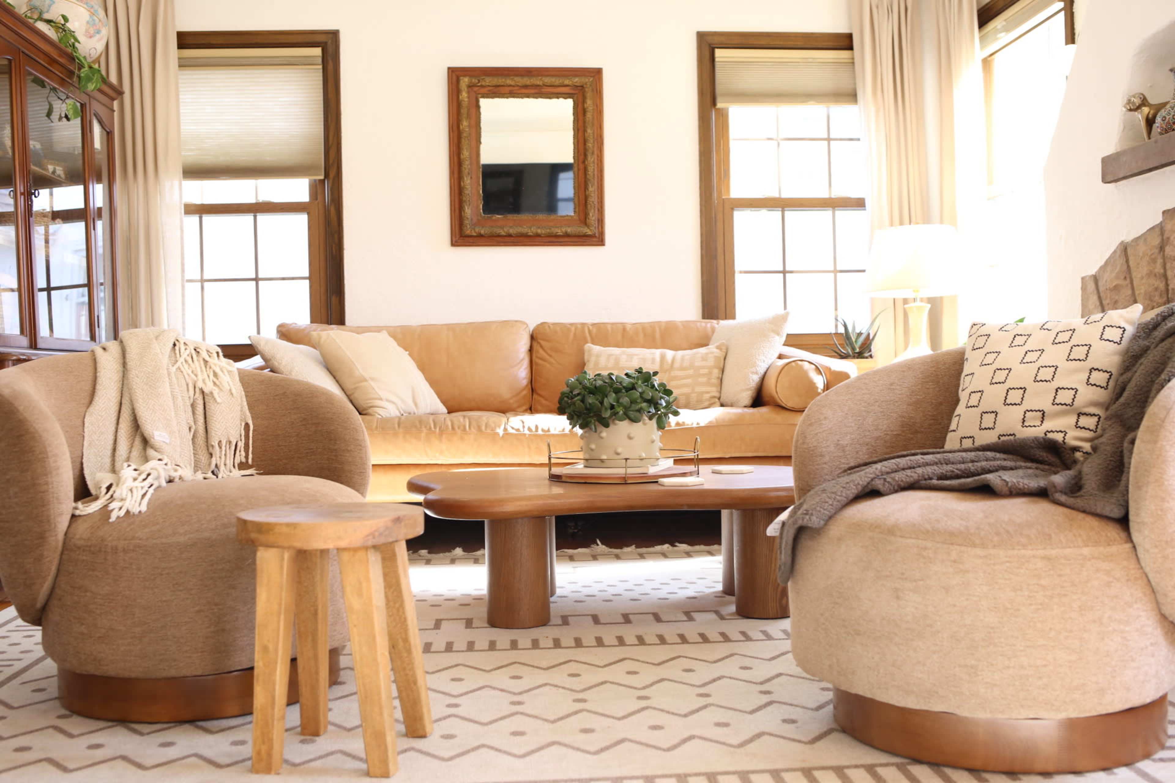The image shows a cozy living room with a beige sofa, two rounded chairs, a wooden coffee table, and large windows allowing natural light.