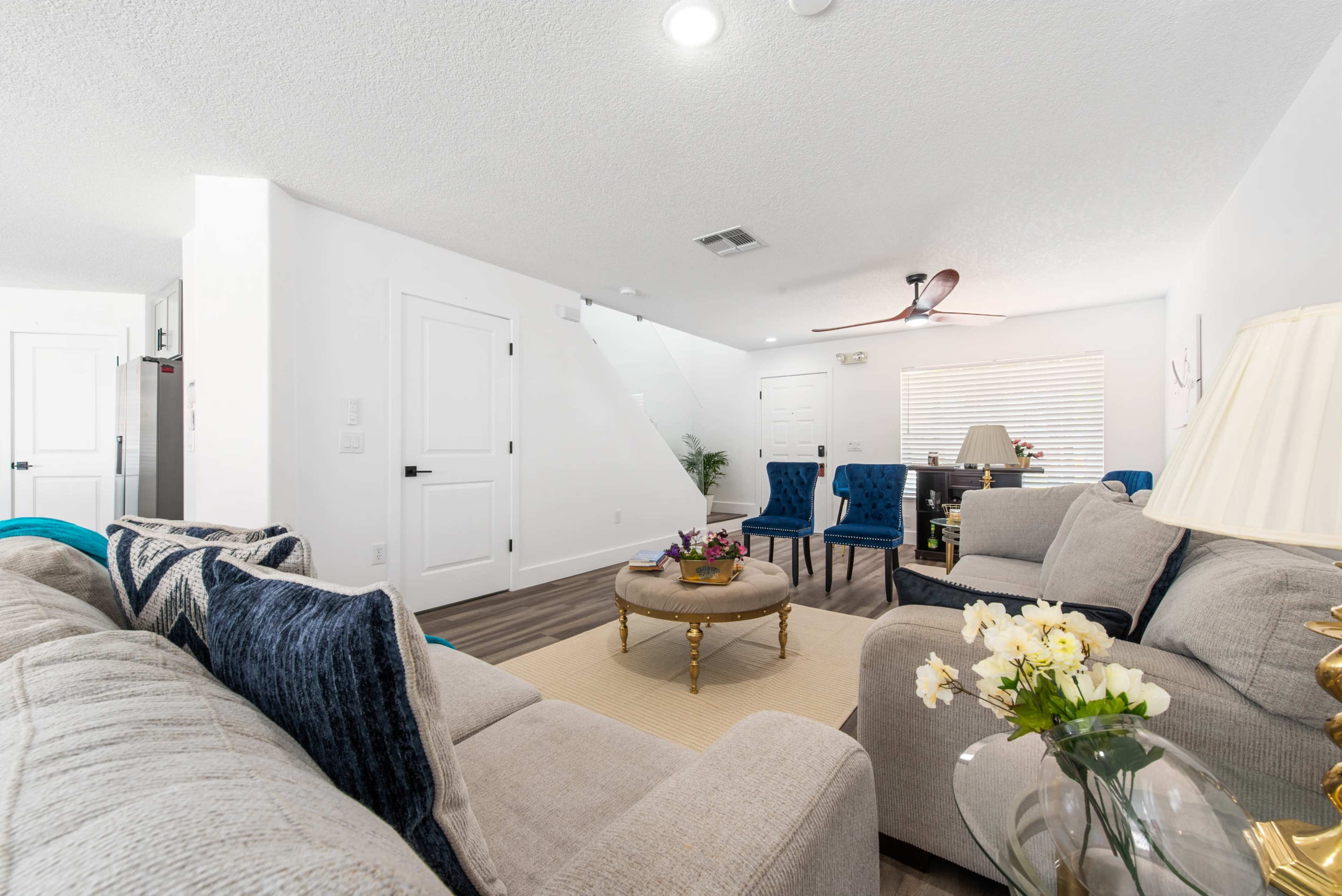 A modern living room with neutral-colored sofas, a round coffee table, and blue upholstered chairs, featuring a ceiling fan and natural light from a window.