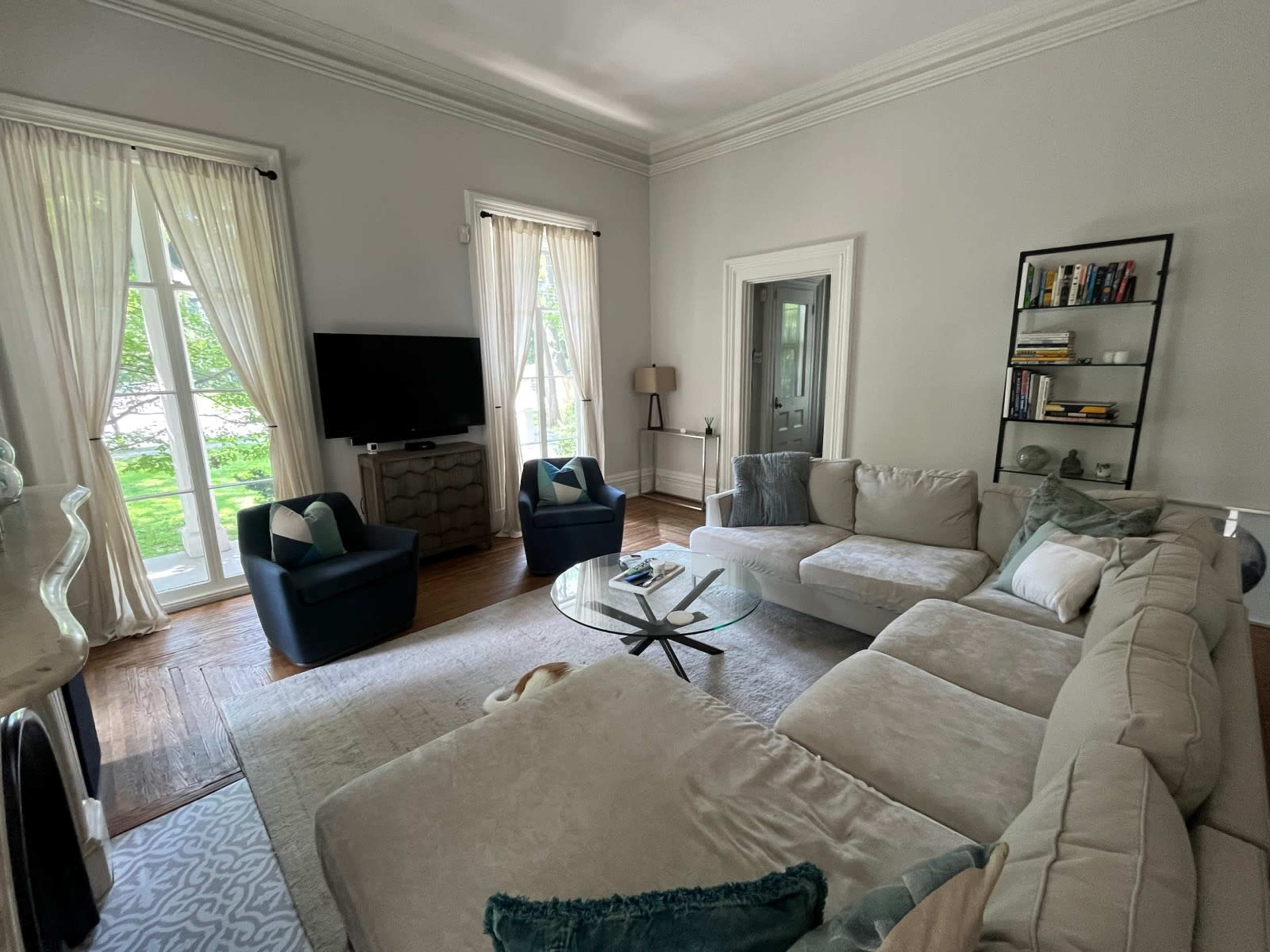 The image depicts a bright living room featuring a large sectional sofa, two blue armchairs, a glass coffee table, and large windows with white curtains.