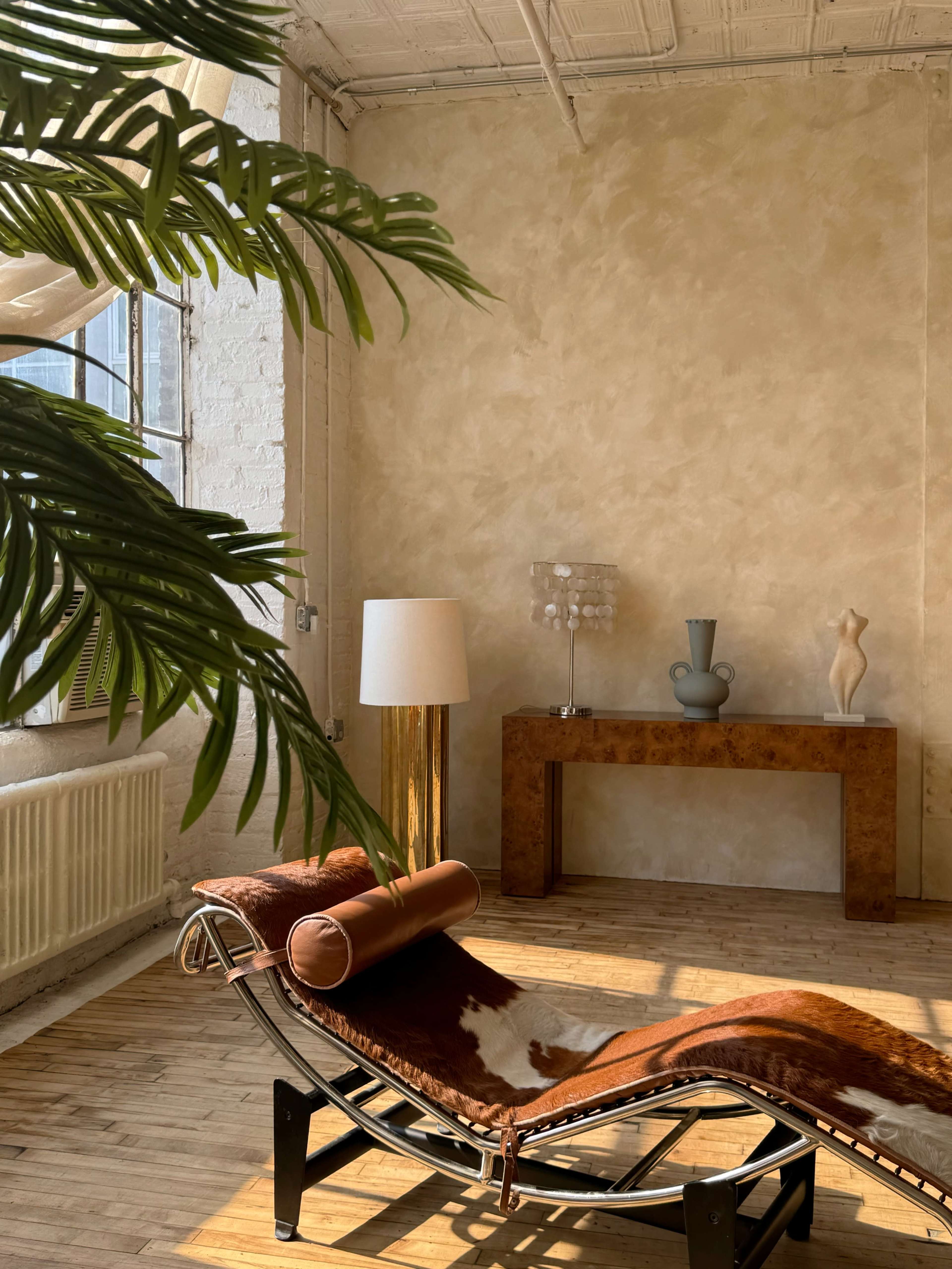 The image features a minimalist interior with a chaise lounge, a lamp, a sculpture, and a console table against a textured wall.