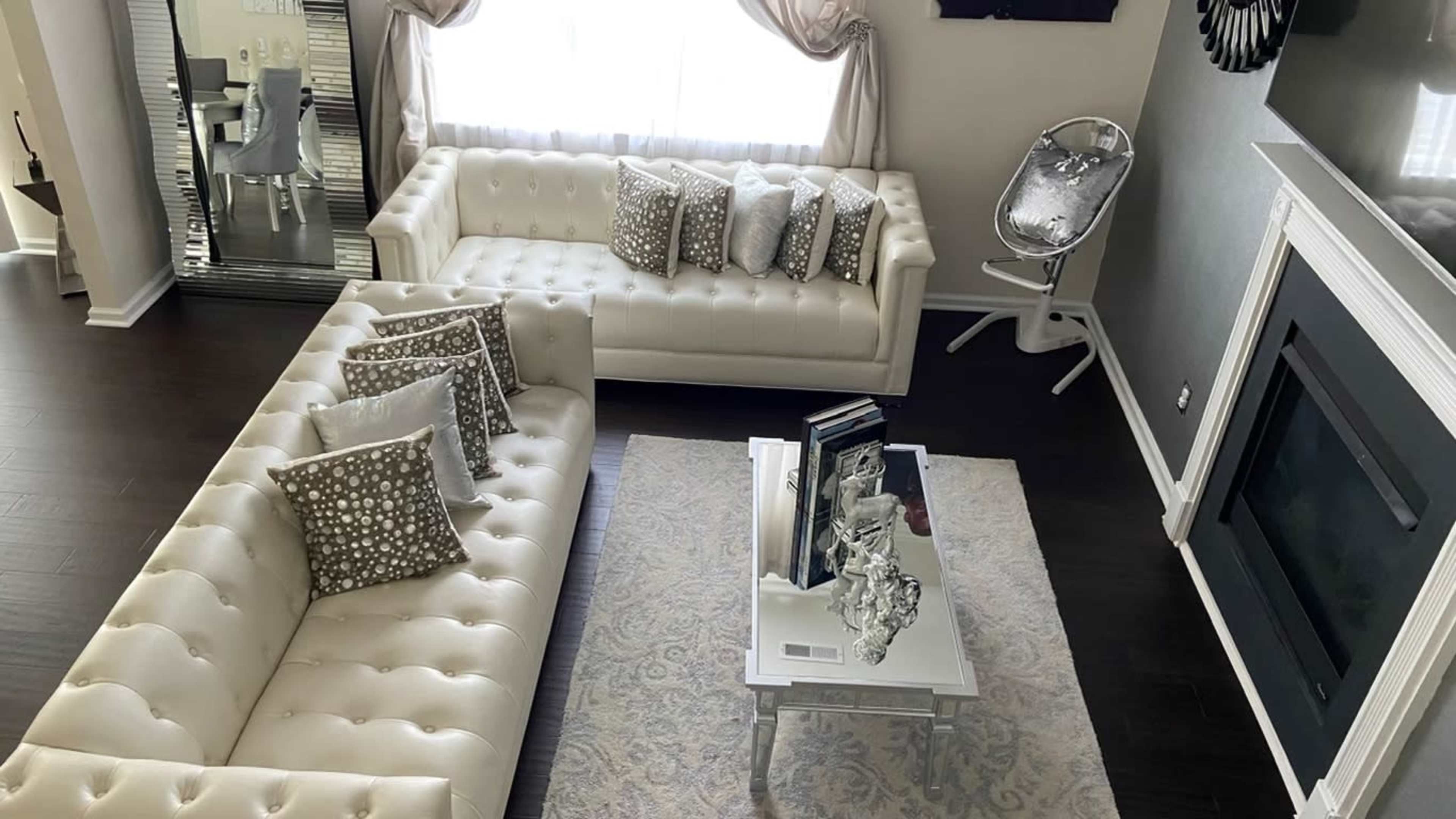 A living room featuring two cream-colored sofas arranged around a coffee table with decorative items, a large window with sheer curtains, and a fireplace.