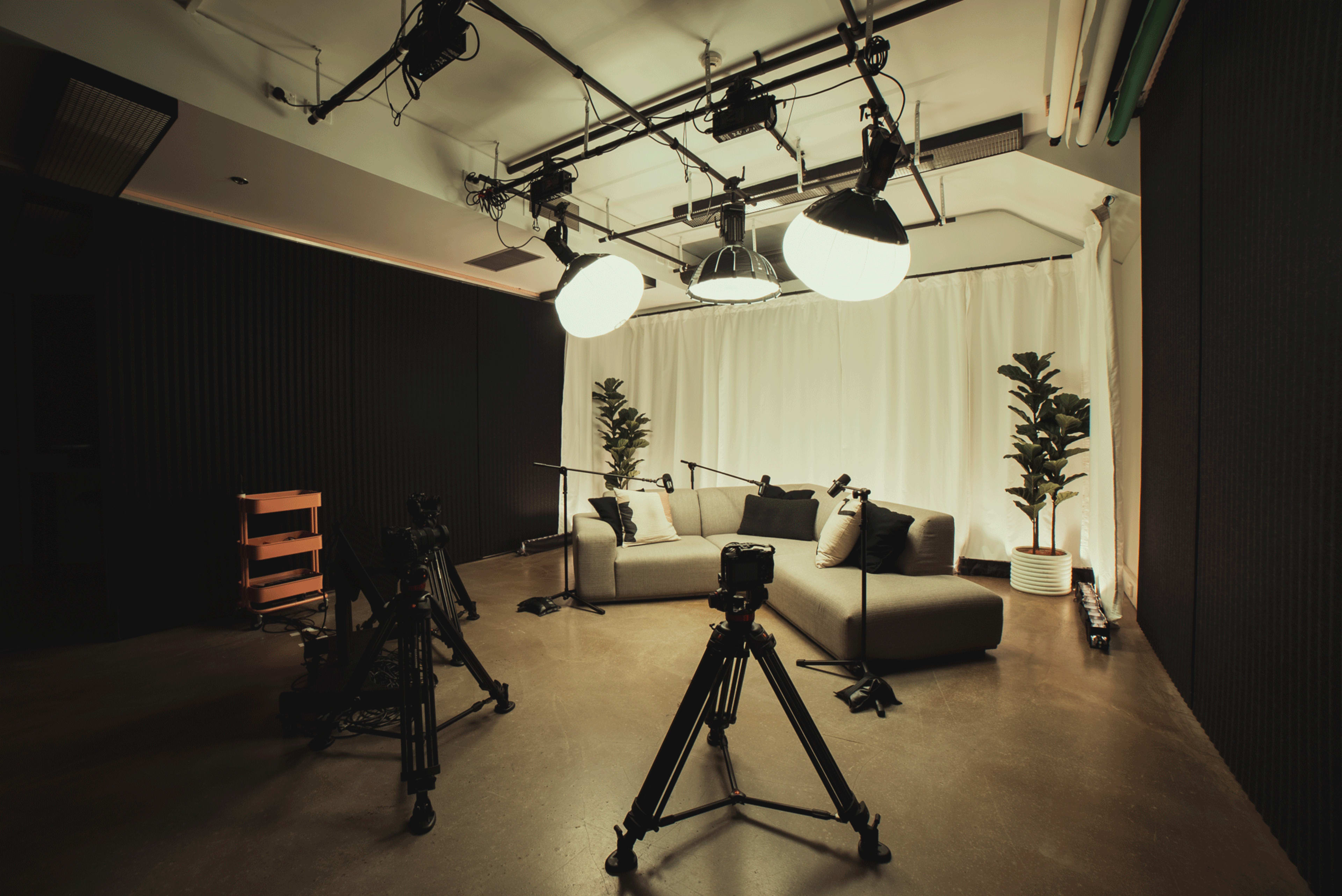 A multimedia studio is set up with two sofas, a white backdrop, and three cameras on tripods.