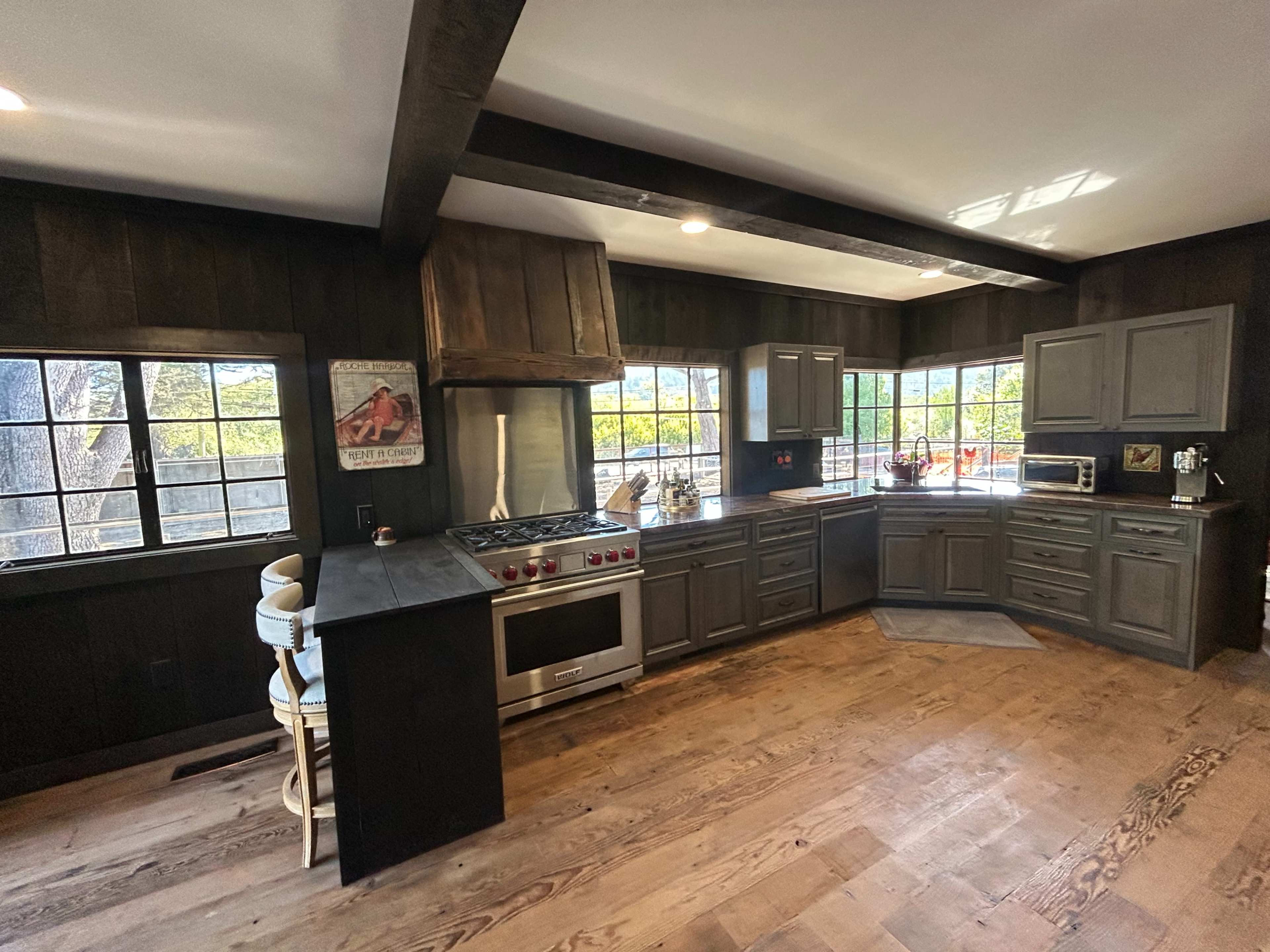 The image shows a modern kitchen with dark wooden cabinetry, a stainless steel oven, and large windows providing natural light.