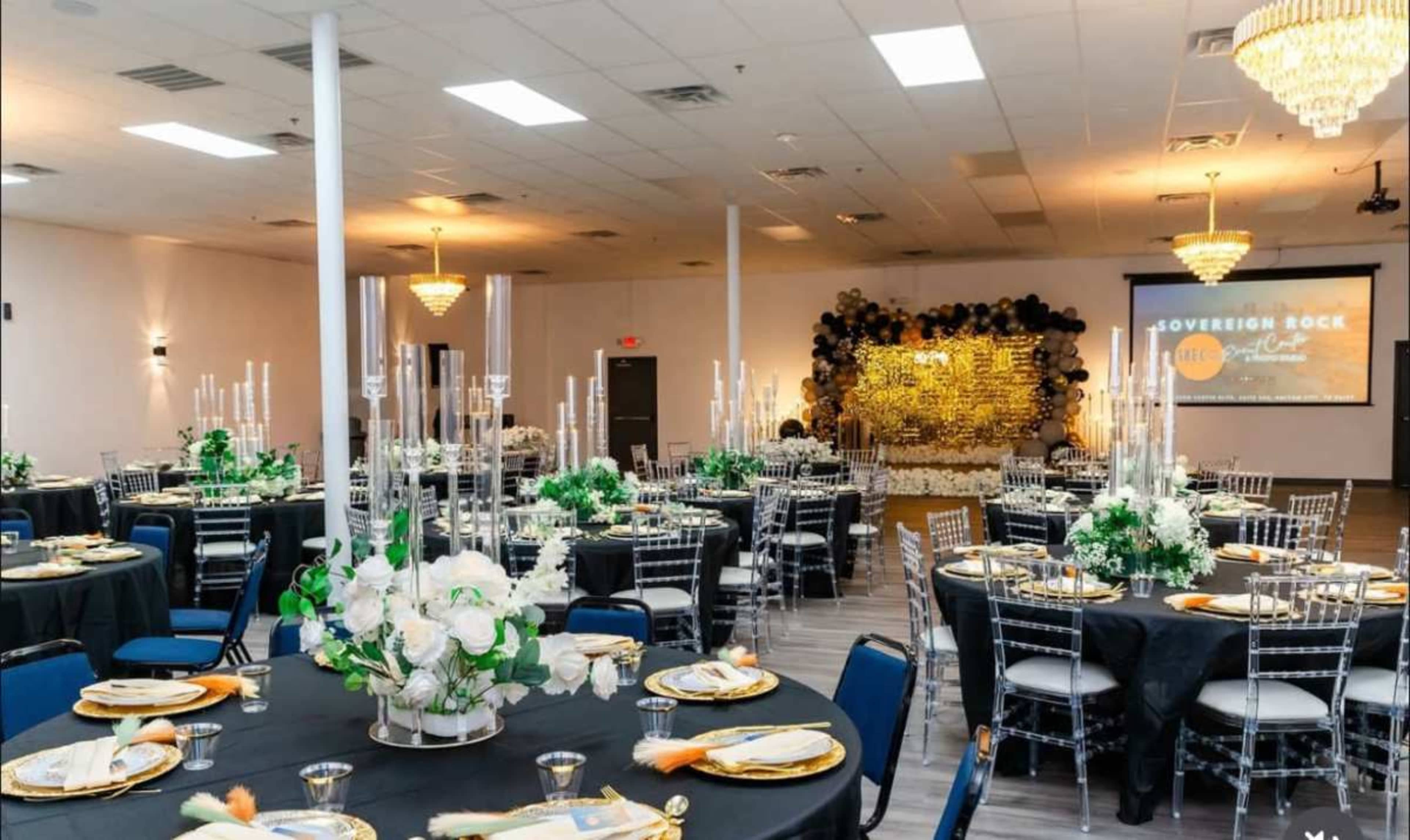 The image shows a decorated banquet hall set up for an event, featuring tables with black linens, gold accents, and floral centerpieces, along with a backdrop of black and gold decorations.