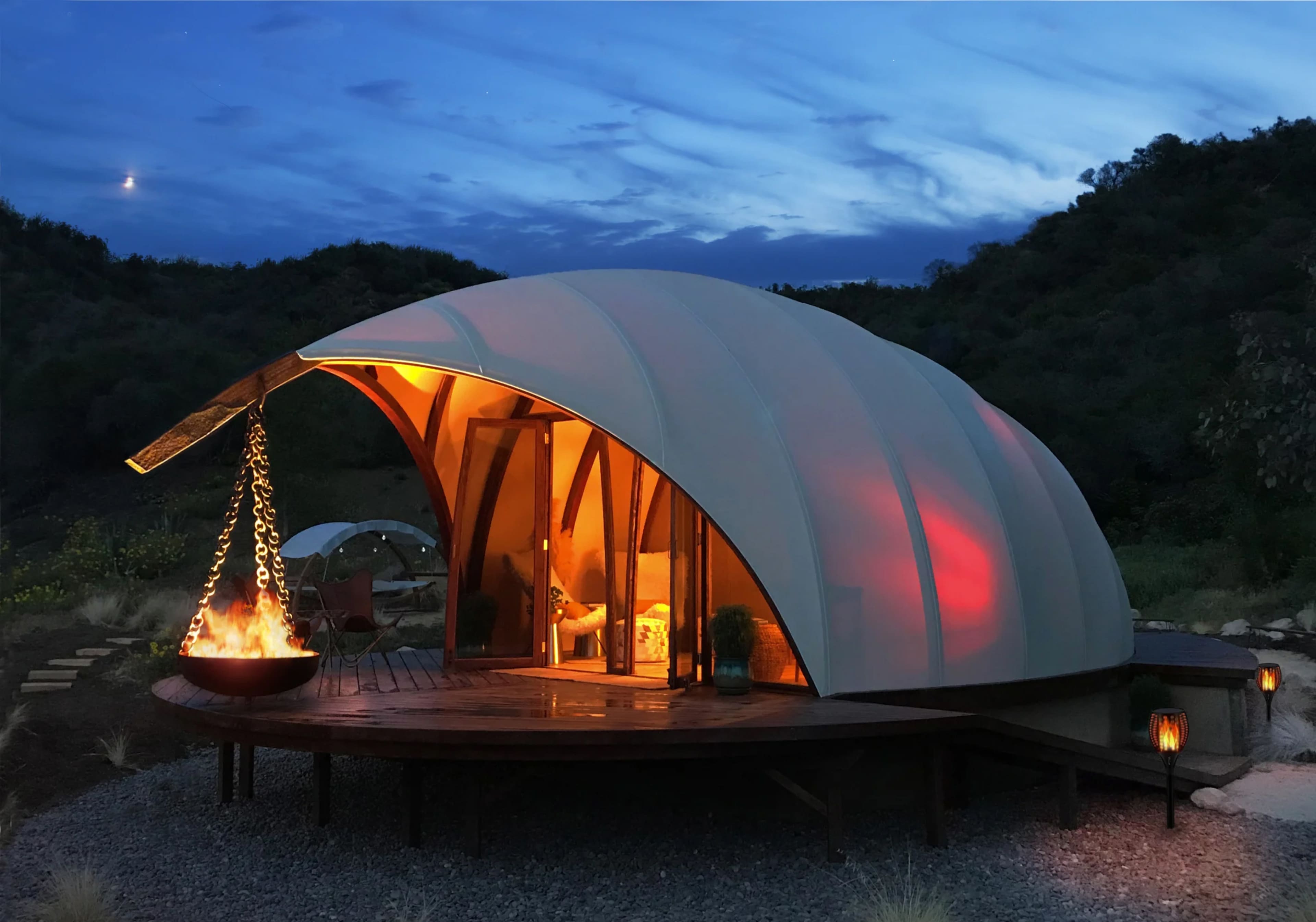 A modern, dome-shaped structure with glowing interiors is set on a wooden deck, surrounded by a natural landscape under a twilight sky.