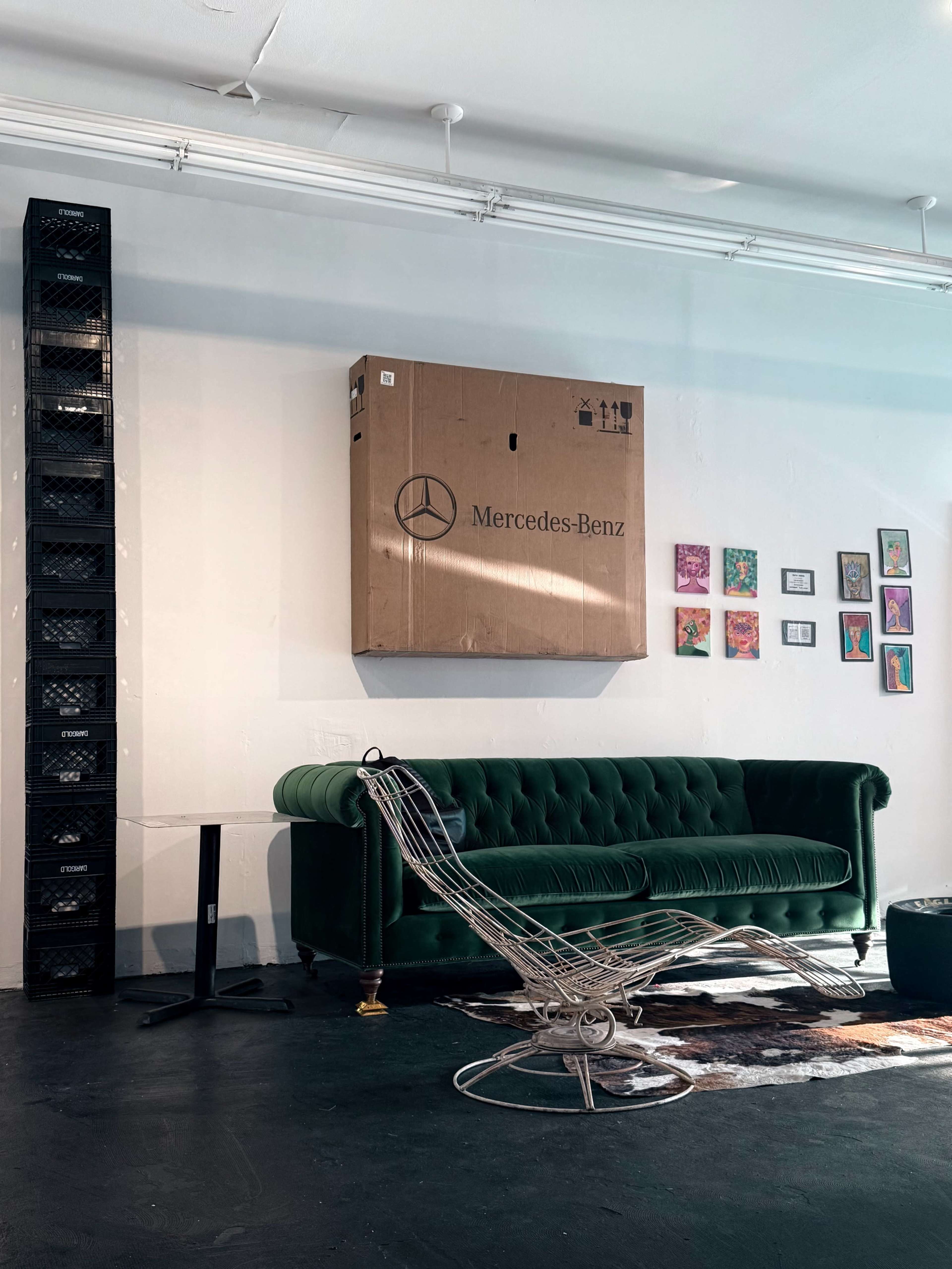 A green velvet sofa is positioned in front of a wall adorned with framed artwork, while a large cardboard box labeled "Mercedes-Benz" hangs above it.