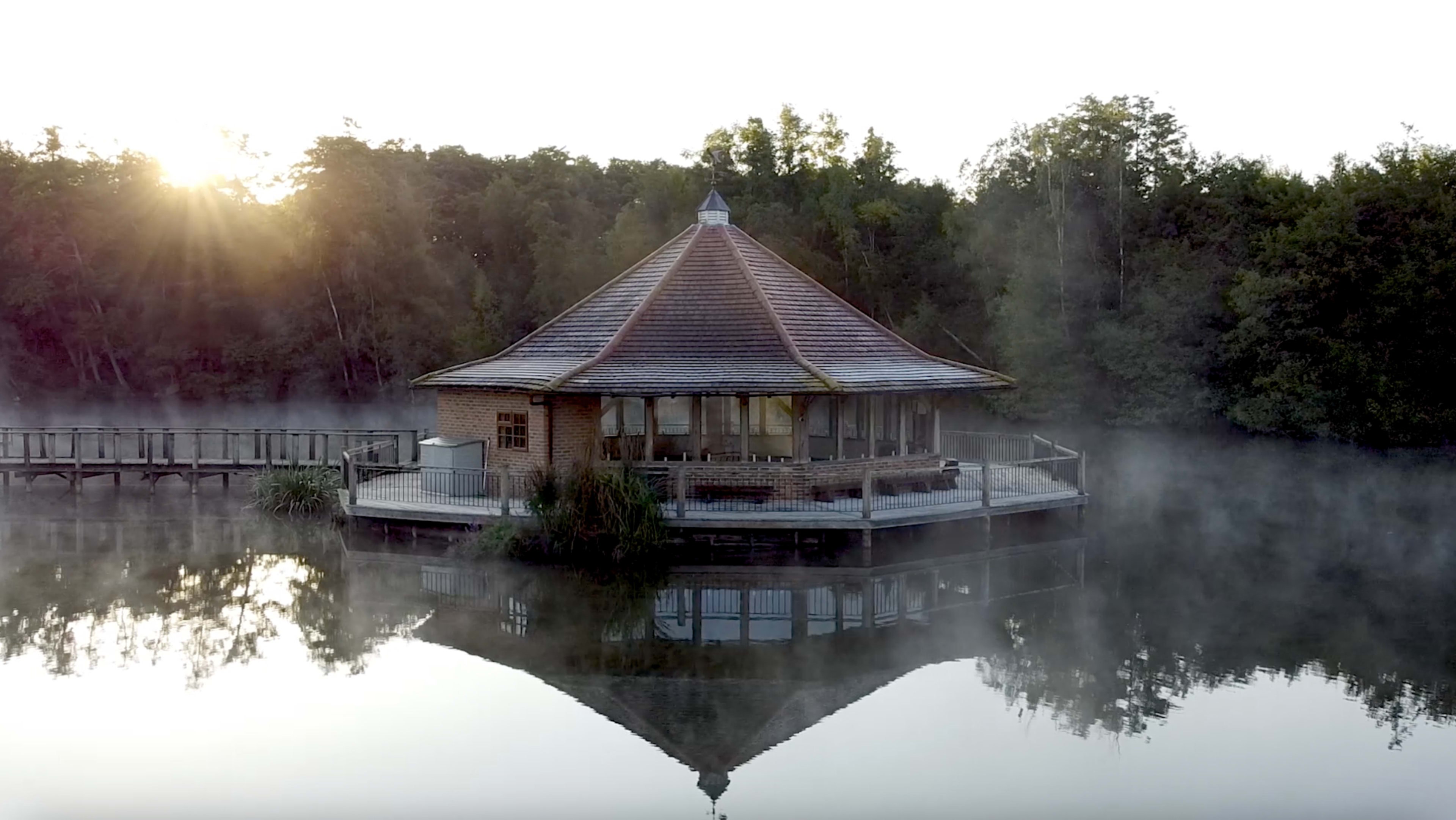 A circular wooden pavilion sits on stilts over a misty lake, surrounded by trees, reflecting in the still water.