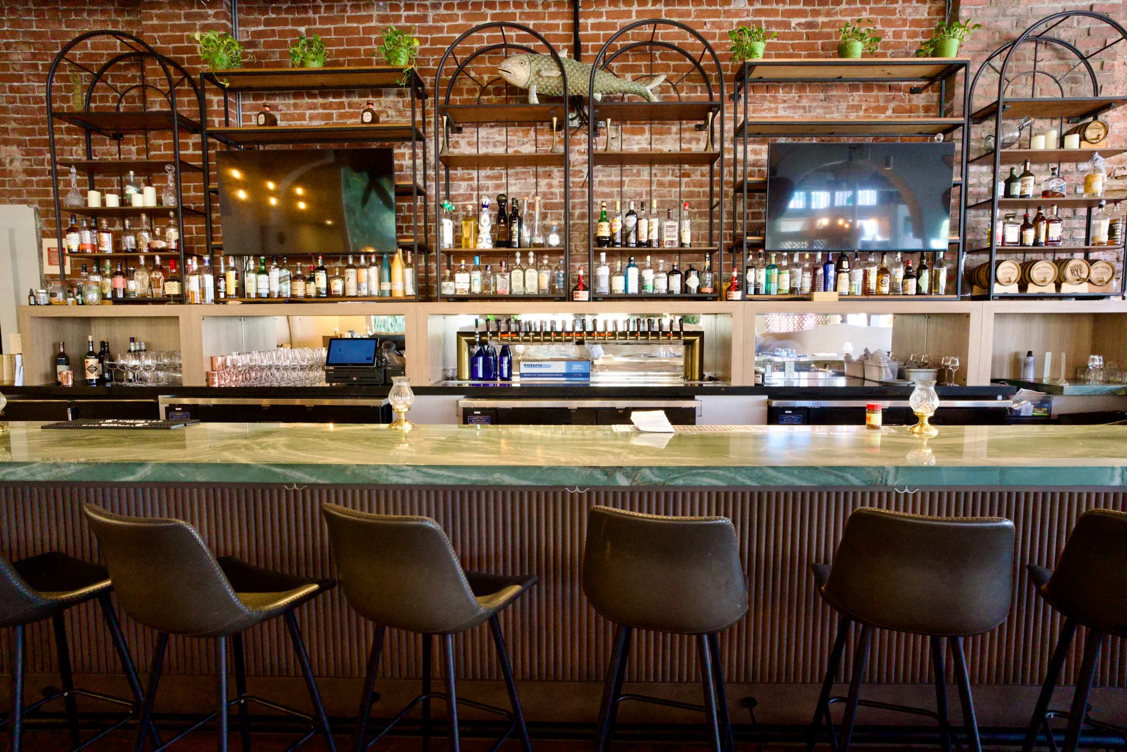 A modern bar with a marble countertop, high chairs, and a background of shelves displaying various bottles and plants against a brick wall.