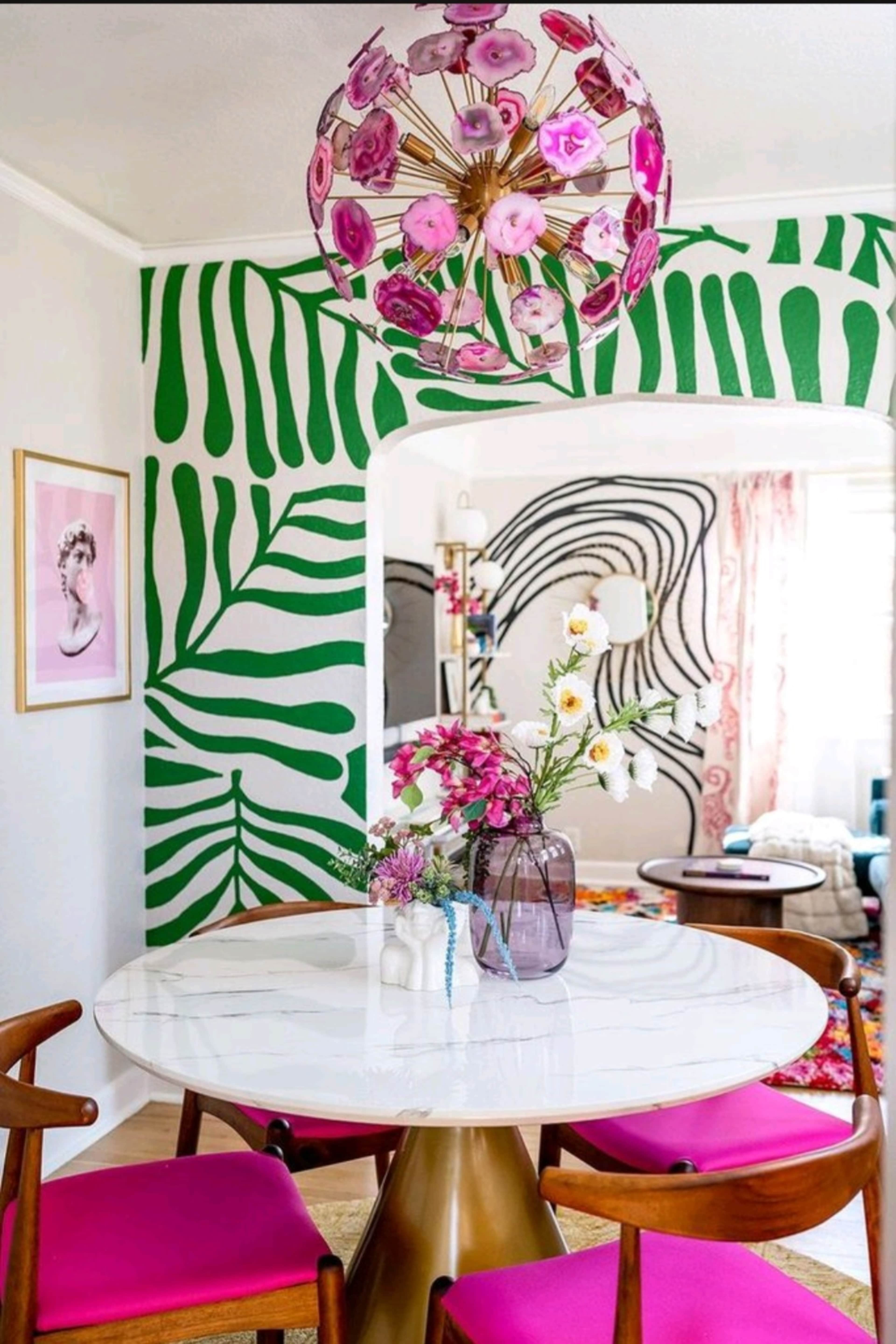 A round dining table with pink chairs is centered in a room featuring bold green leaf-patterned walls and a colorful chandelier.