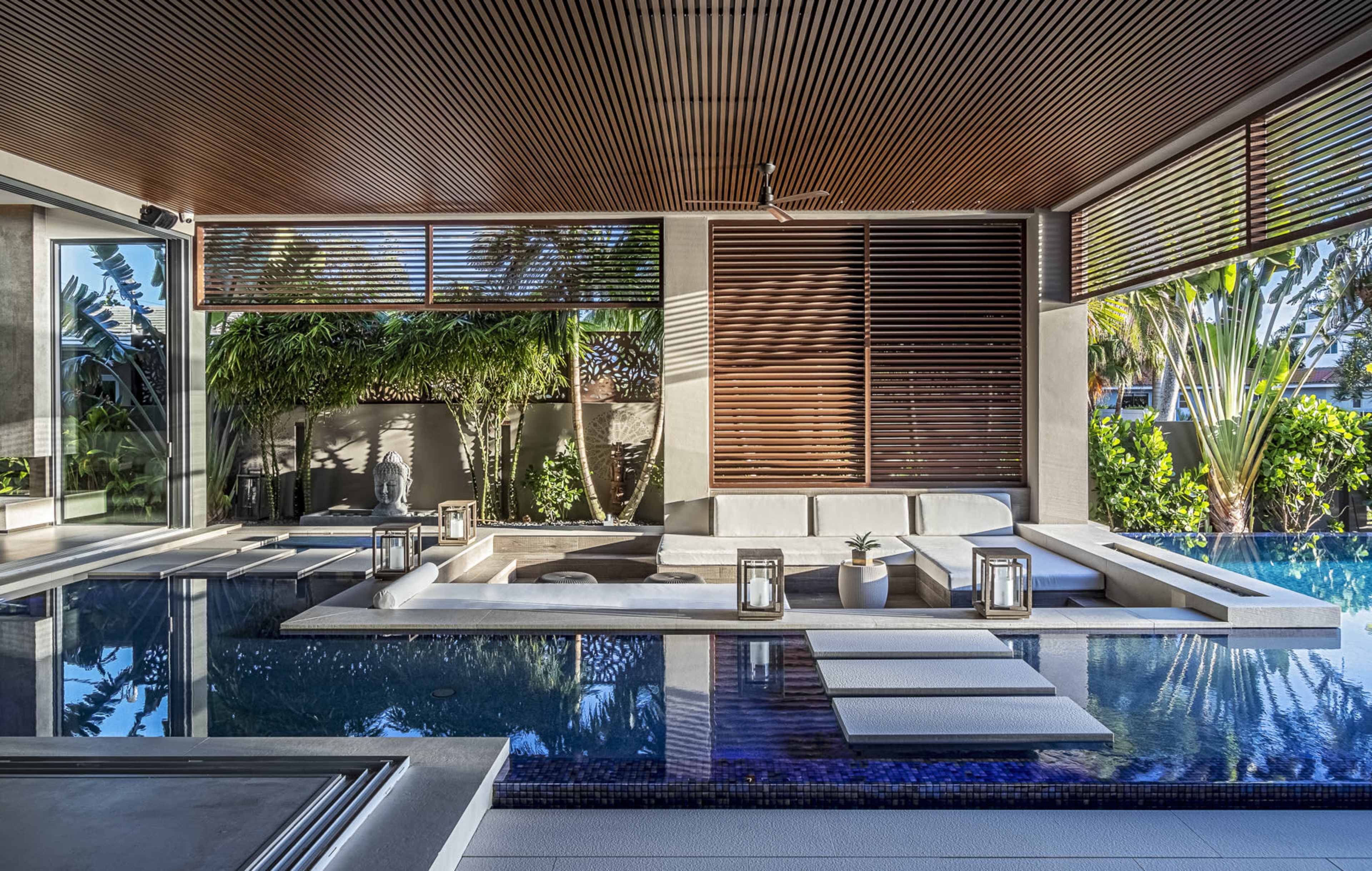 A modern living space featuring a pool area with a sofa, lanterns, and tropical greenery visible through large windows.