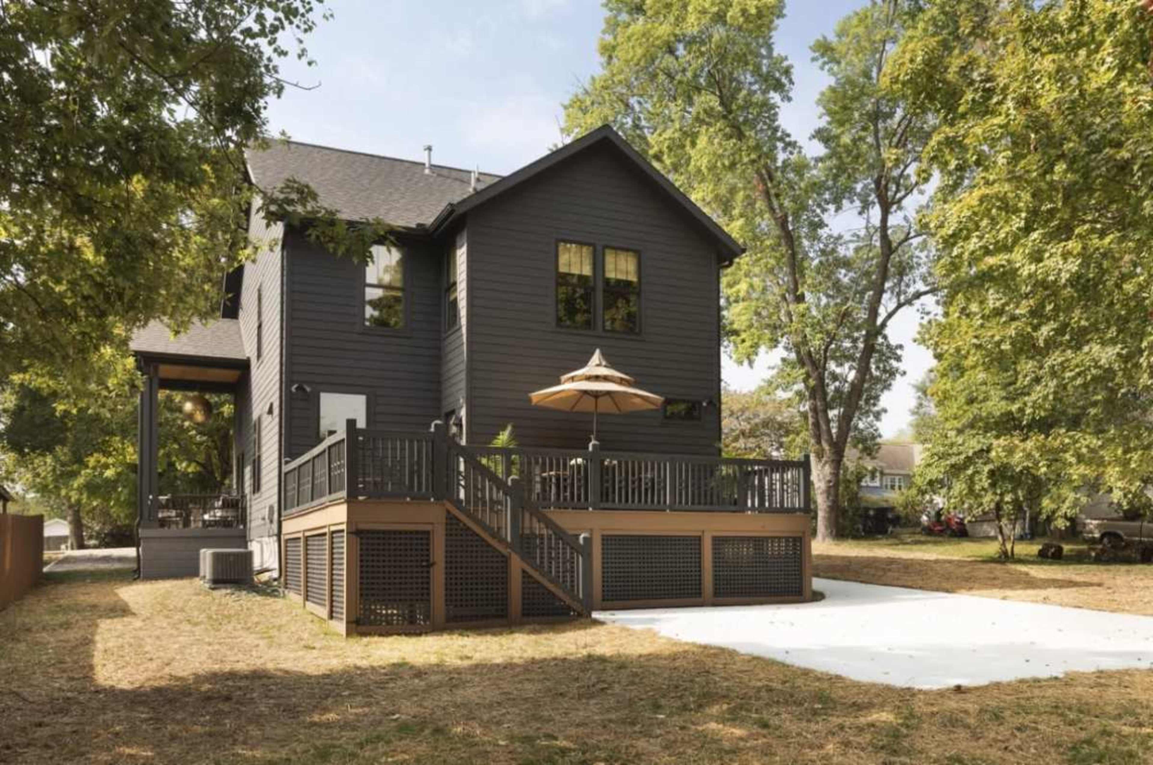 A two-story dark-colored house features a wooden deck with an umbrella, surrounded by trees and a lawn.