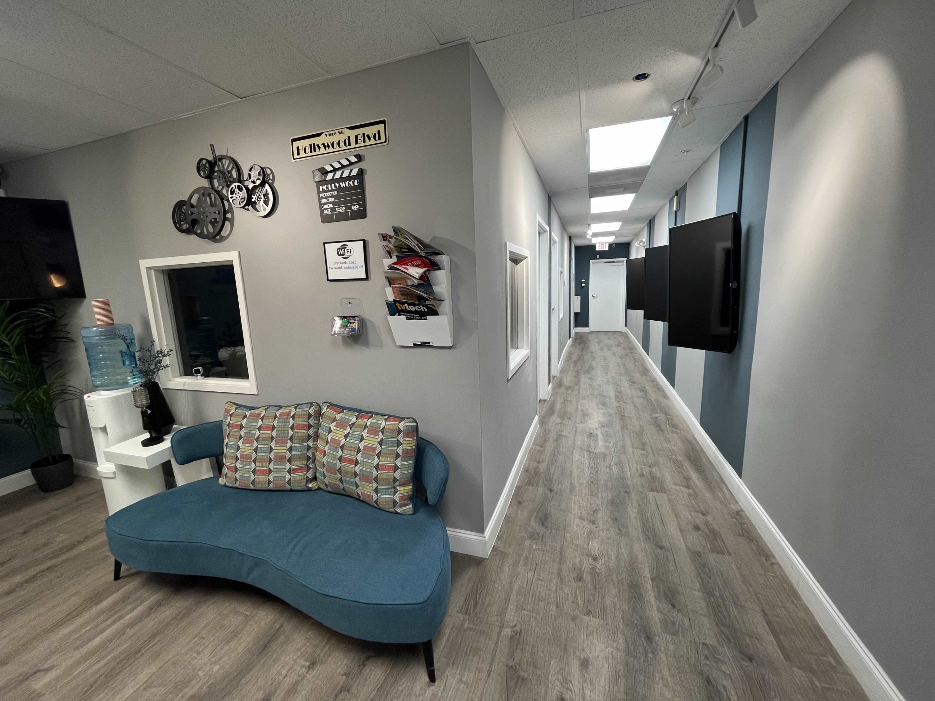 The image shows a modern hallway with a curved blue couch, a water cooler, and several wall decorations, leading to a brightly lit corridor.