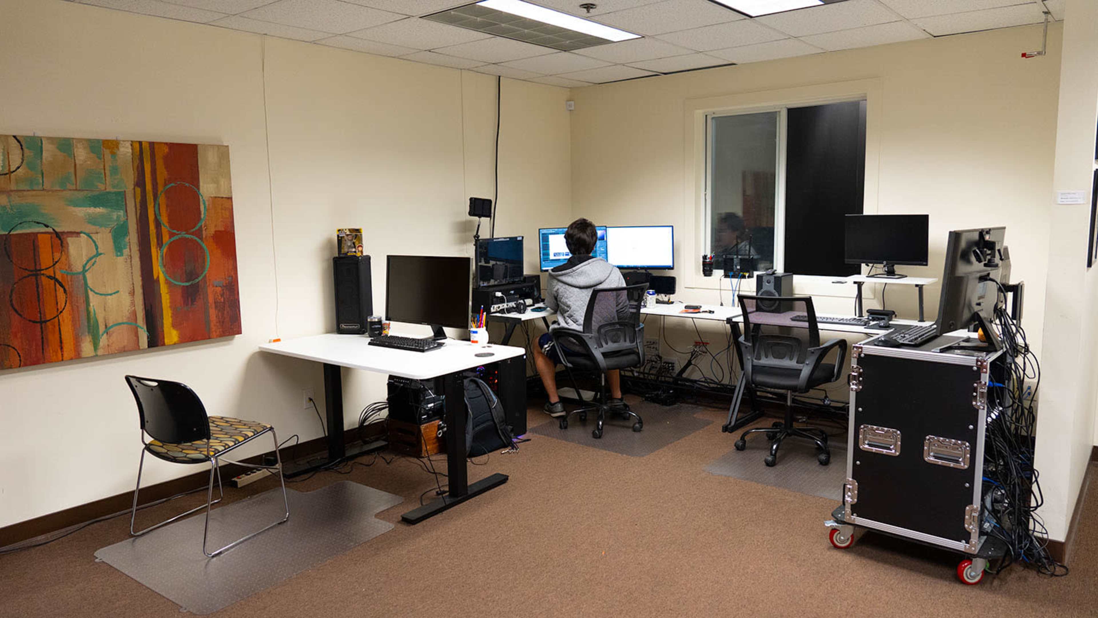 A person is sitting at a desk with multiple monitors in a small office space featuring a colorful painting on the wall.