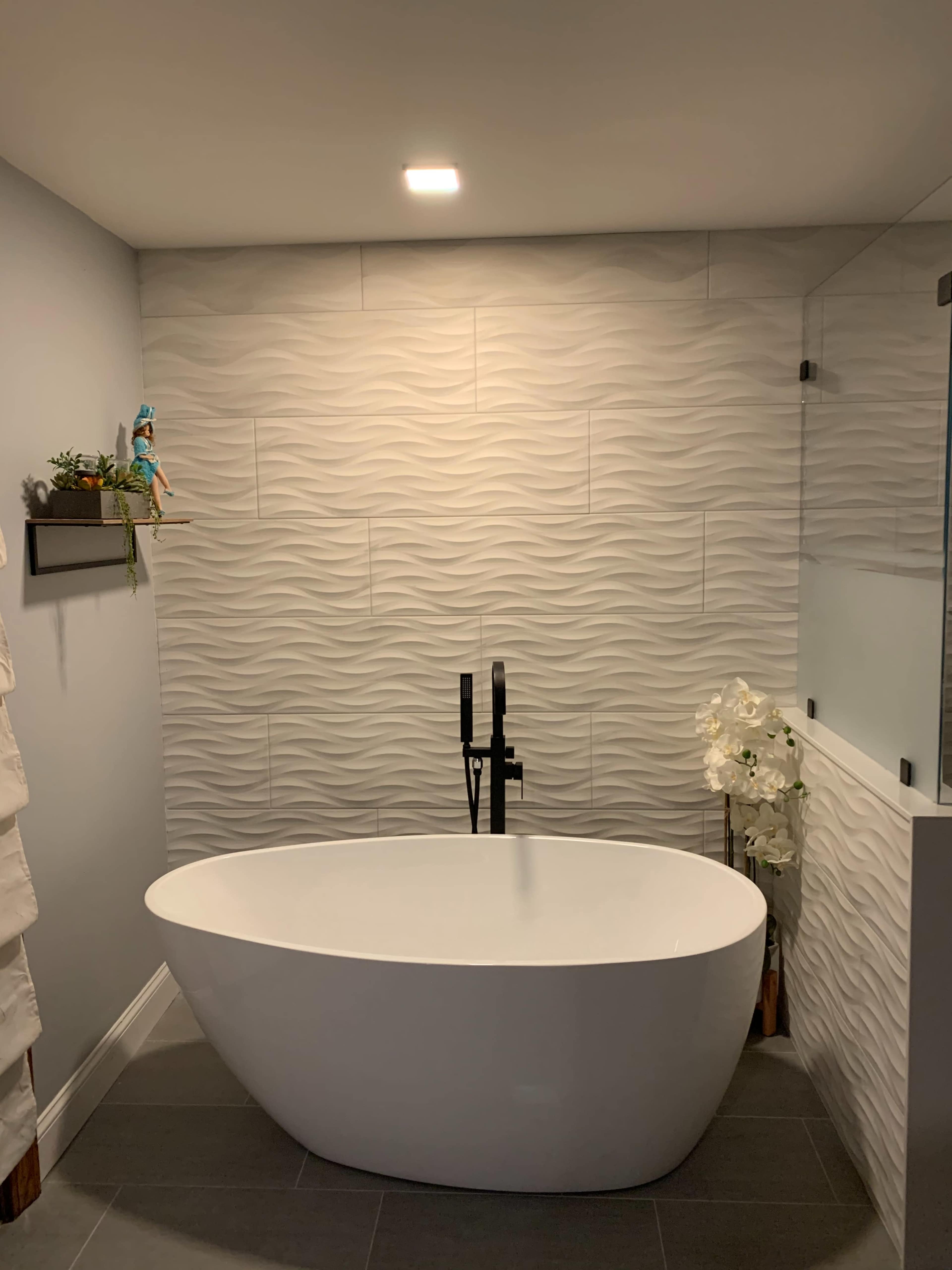 The image shows a modern bathroom featuring a freestanding white bathtub, a textured wave-patterned wall, and a glass shower enclosure.