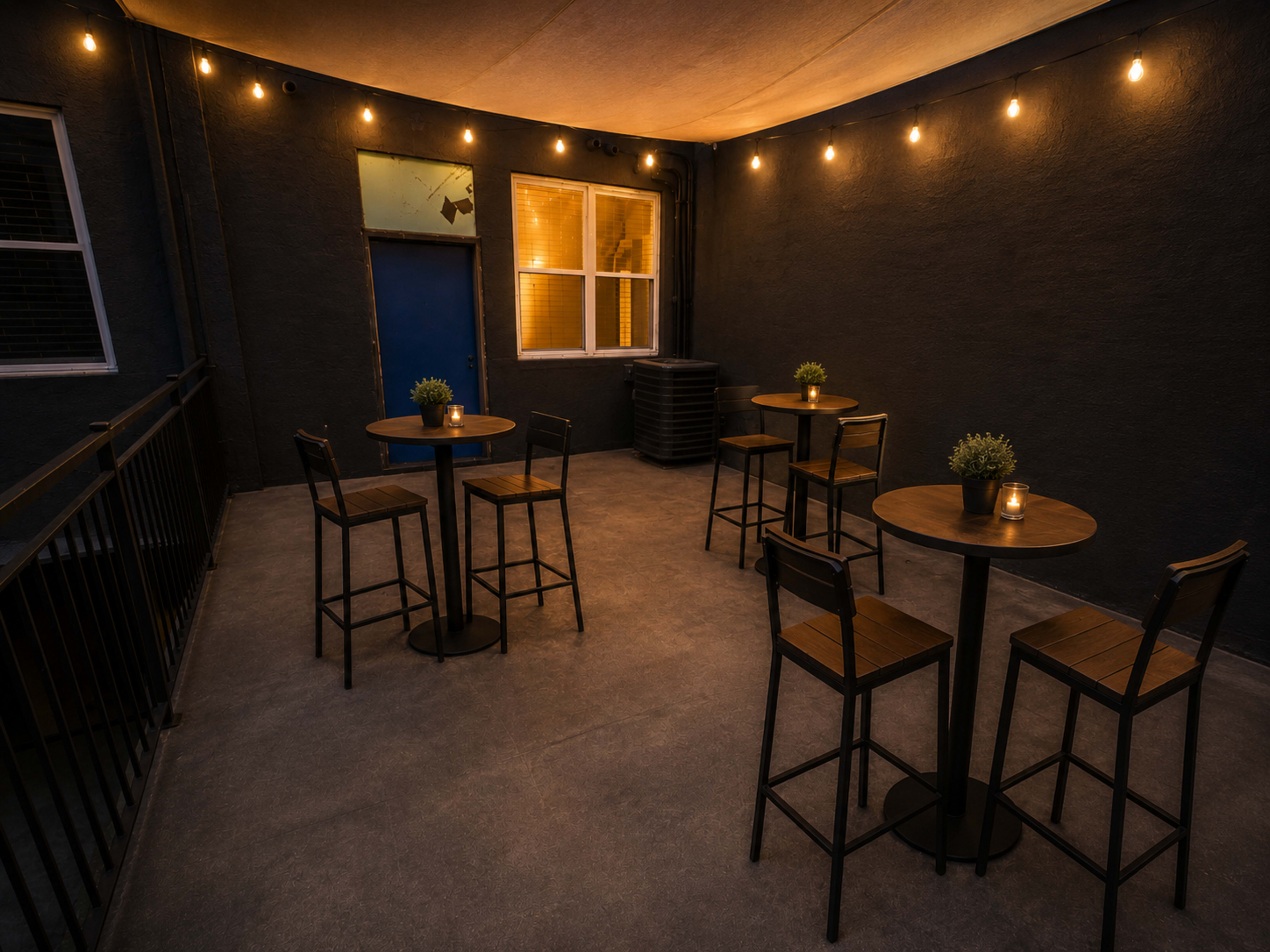 A dimly lit outdoor patio features several tall tables with chairs and small potted plants, creating an intimate setting.