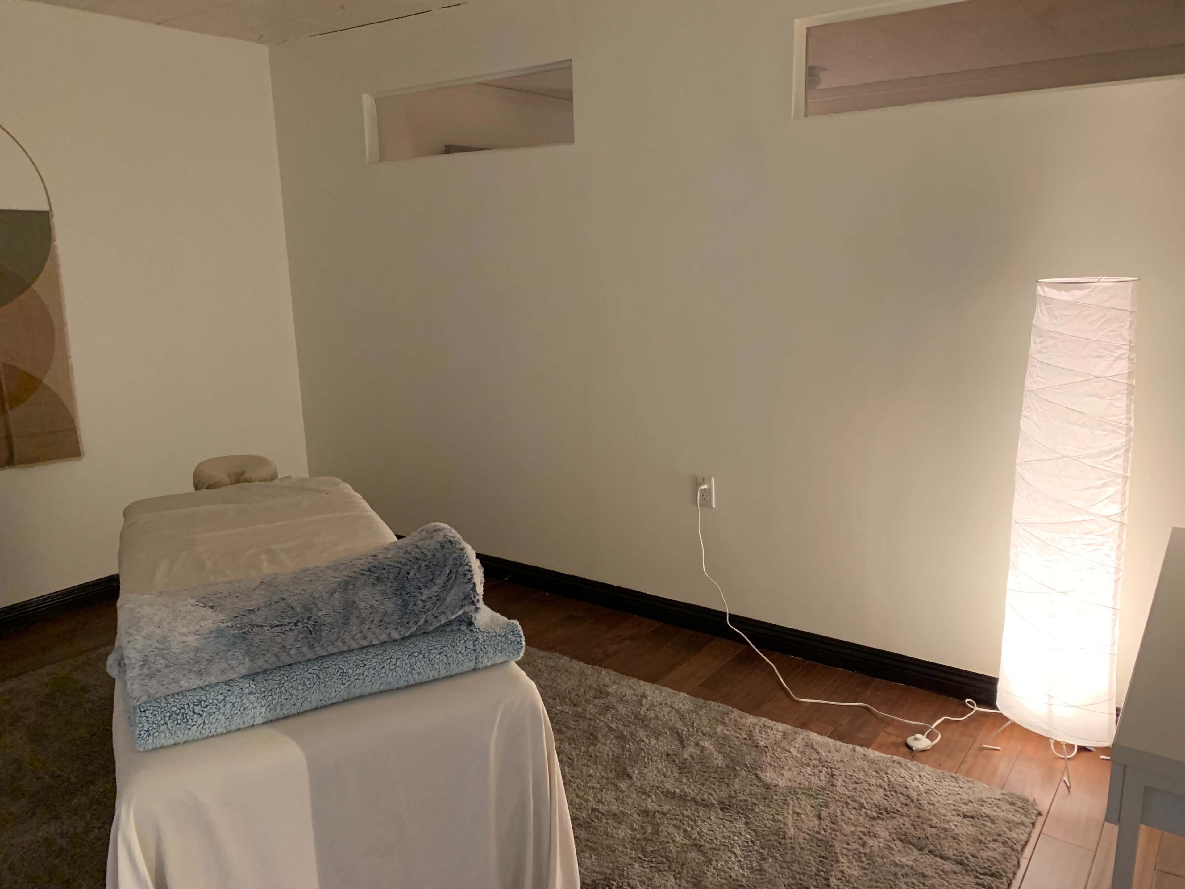 The image shows a minimalist room with a massage table covered in a blue towel and a tall light source illuminating a corner.