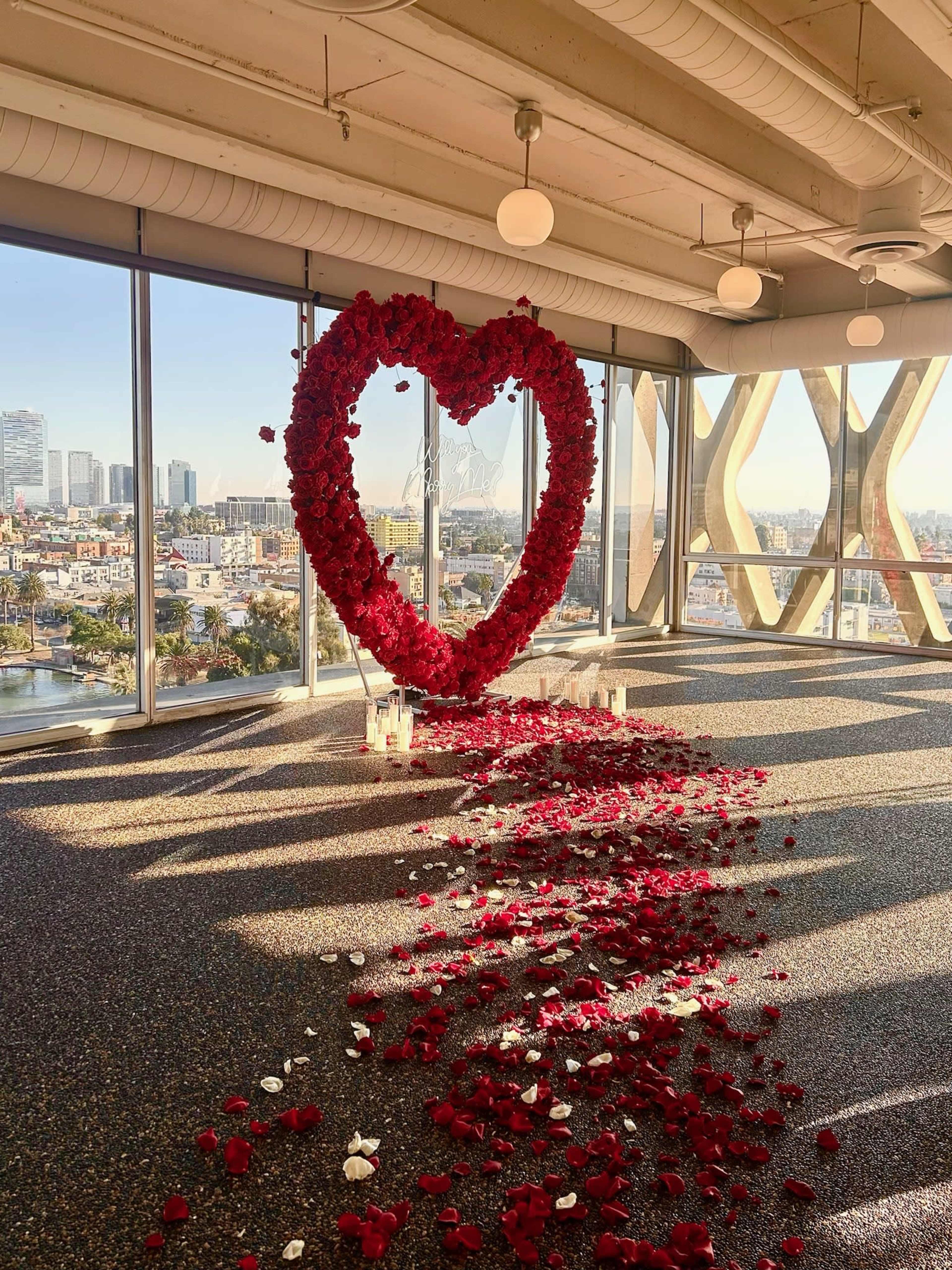 The image shows a large heart-shaped arrangement made of red roses, surrounded by scattered rose petals and candles, set against a city skyline visible through large windows.