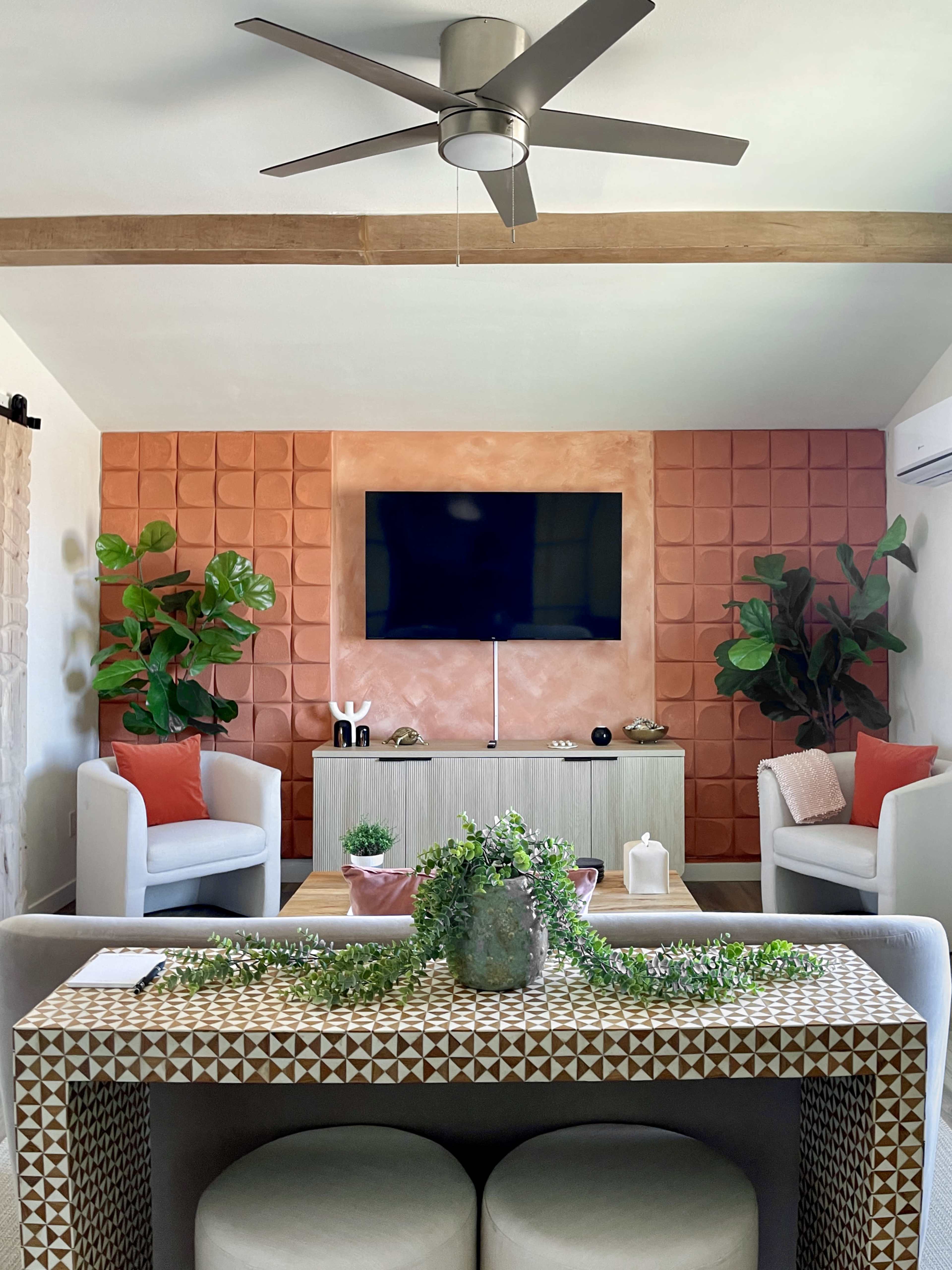 The image shows a modern living room with a television mounted on a textured wall, flanked by potted plants and two armchairs, while a patterned table sits in the foreground.