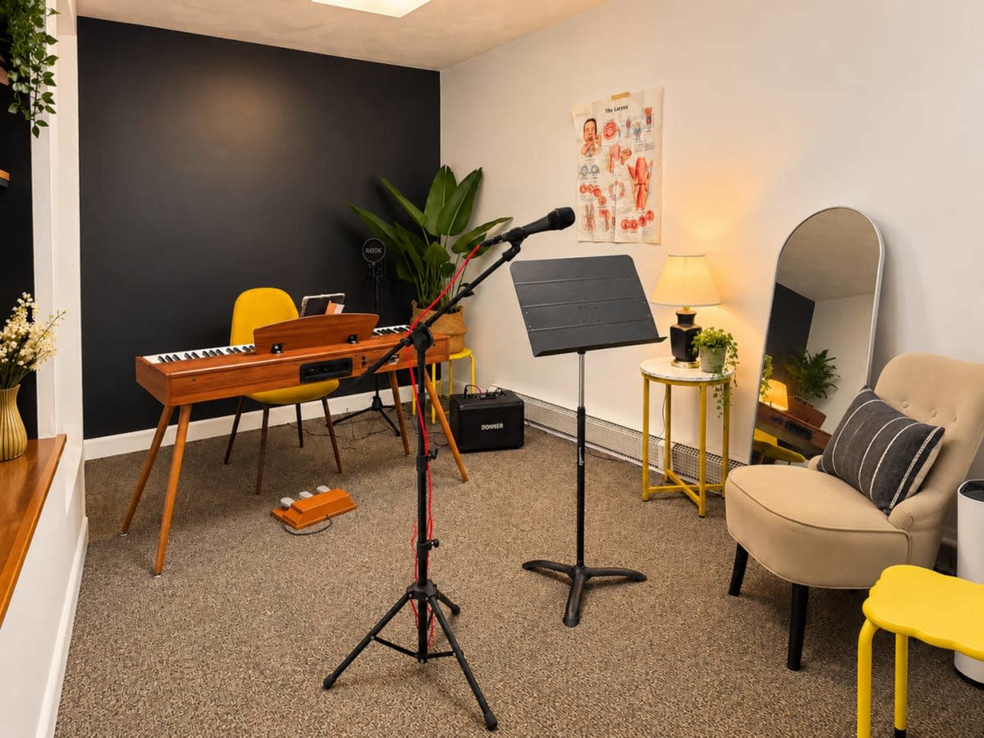 A small music practice room features a keyboard on a stand, a microphone on a stand, a music stand, and a chair, surrounded by plants and decorative elements.