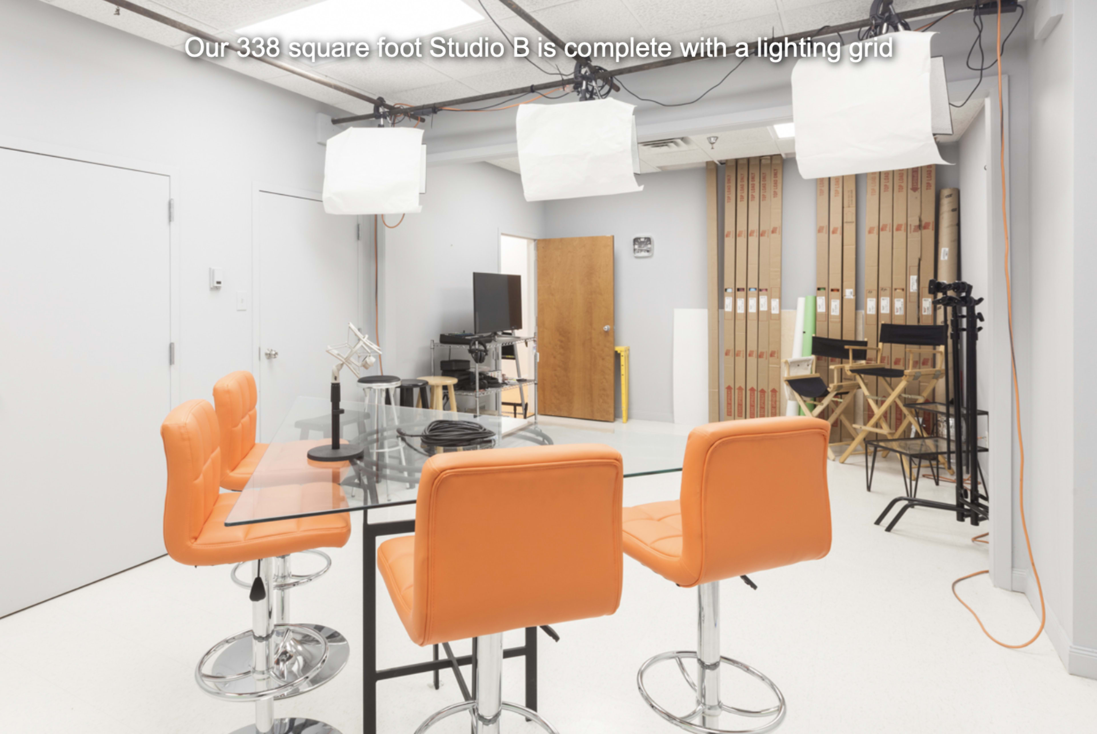 A well-lit studio with orange bar stools around a glass table, equipped with a lighting grid and various studio equipment in the background.