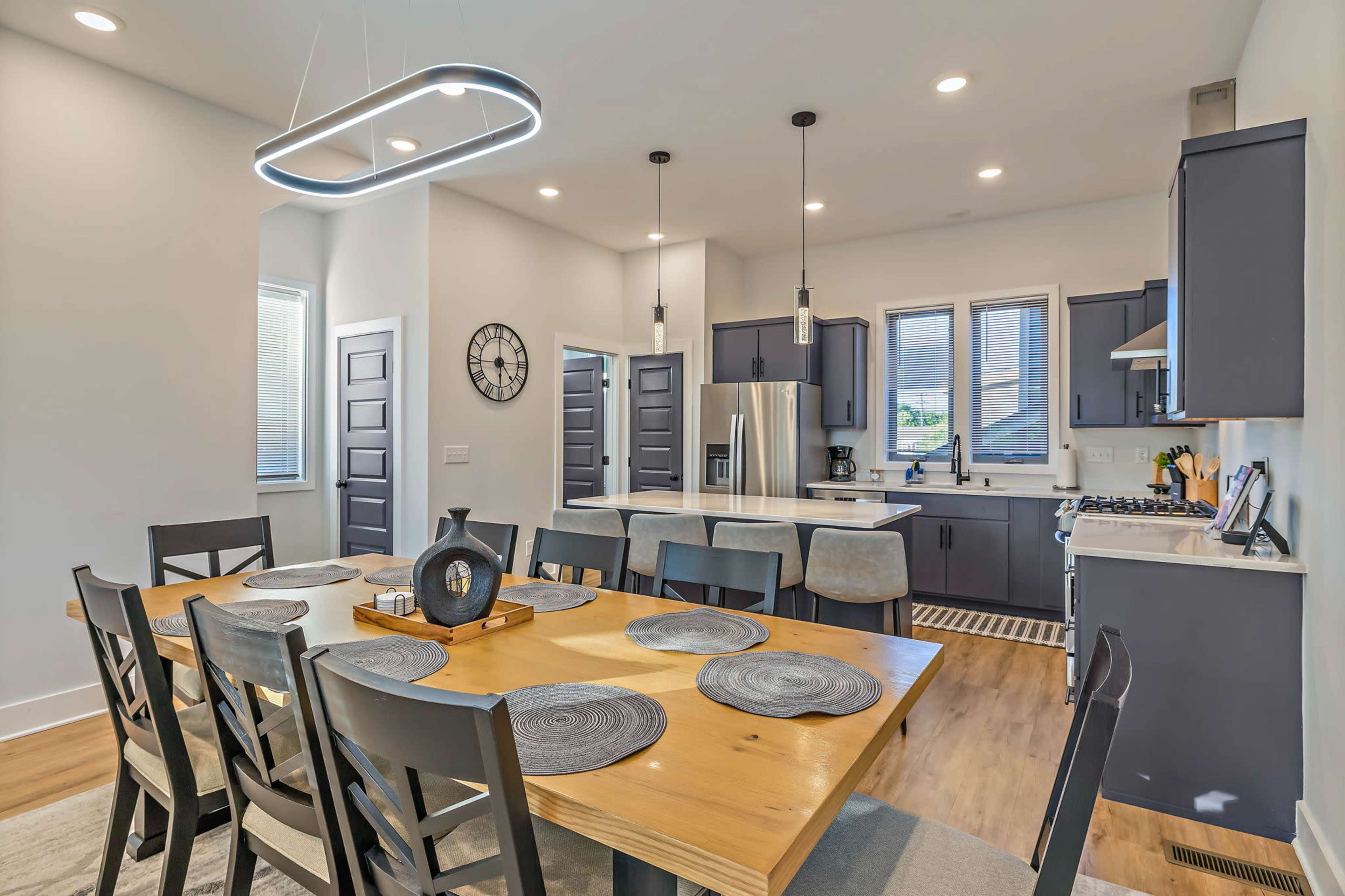 A modern kitchen and dining area features a large wooden table surrounded by black chairs, with a well-equipped kitchen in the background.