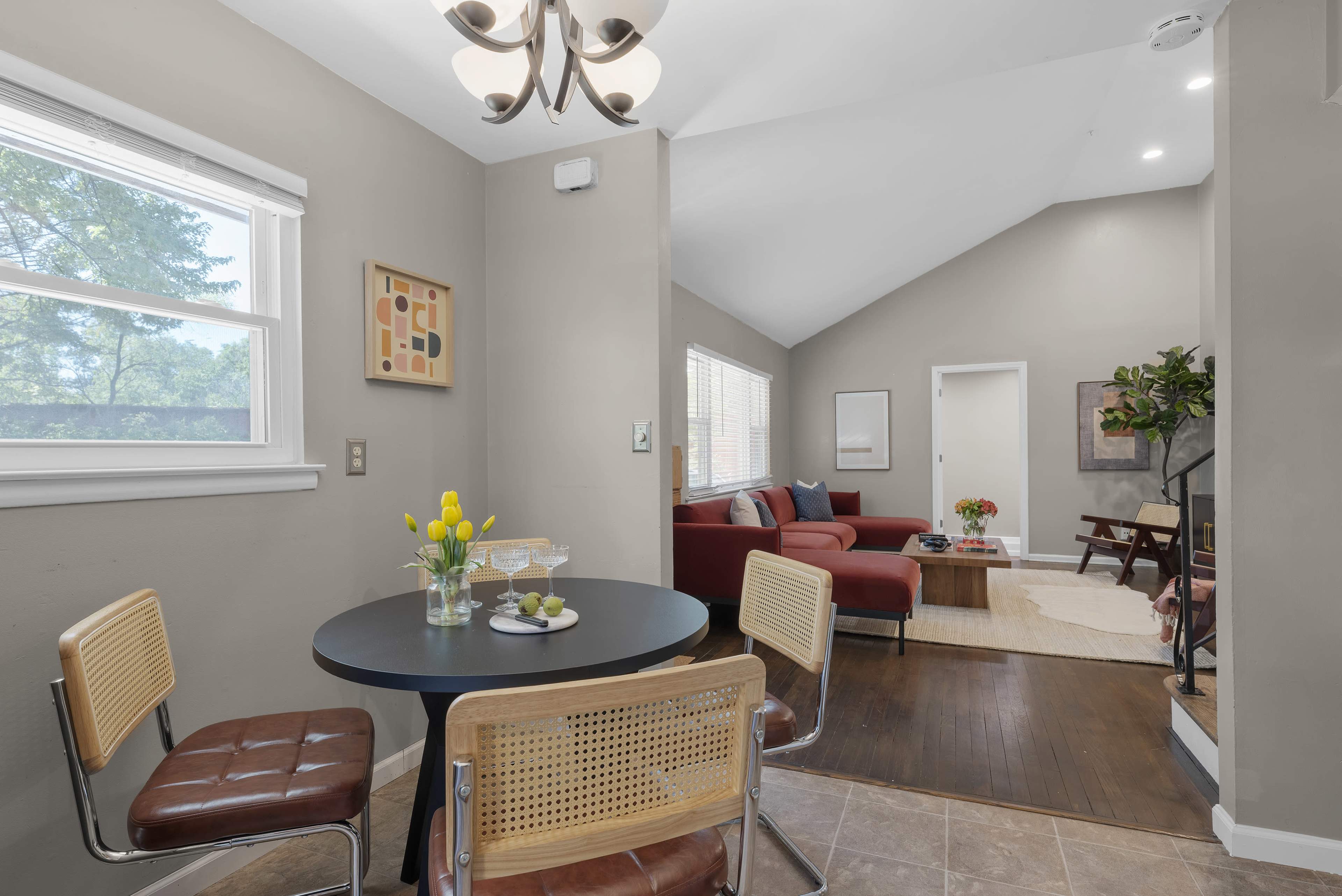 A small dining area with a round black table and two chairs is adjacent to a living room featuring a red sofa and bright natural light from a nearby window.