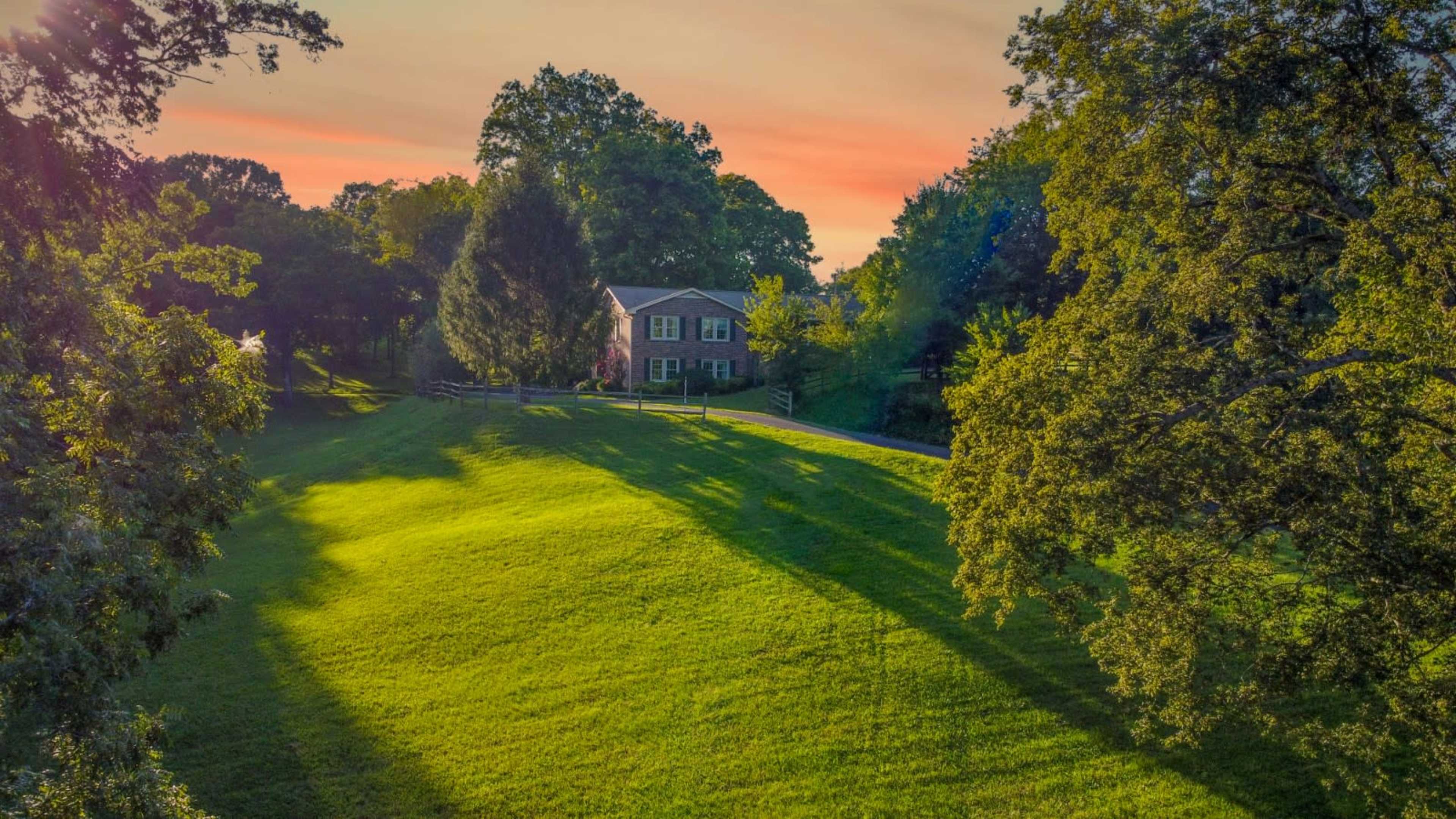 A two-story brick house is situated on a gently sloping green lawn under a colorful sunset sky.