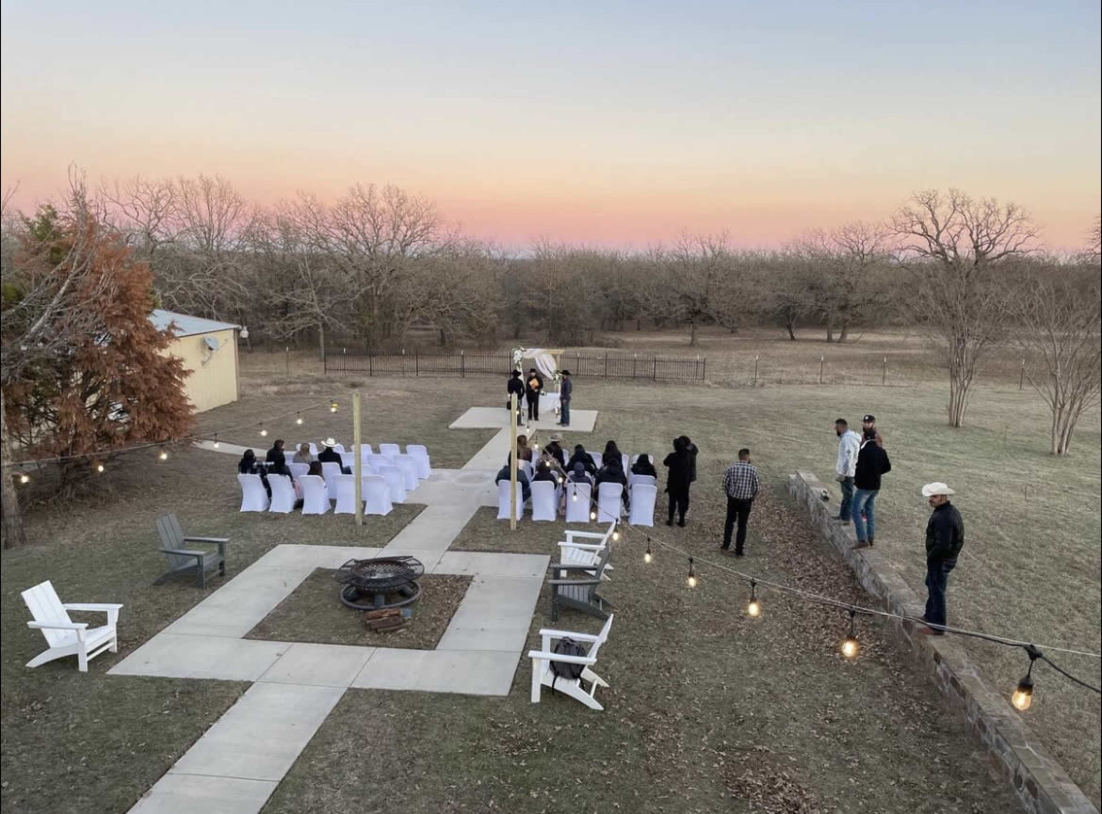 A wedding ceremony is set up outdoors with chairs arranged facing an altar, surrounded by trees and a sunset sky.