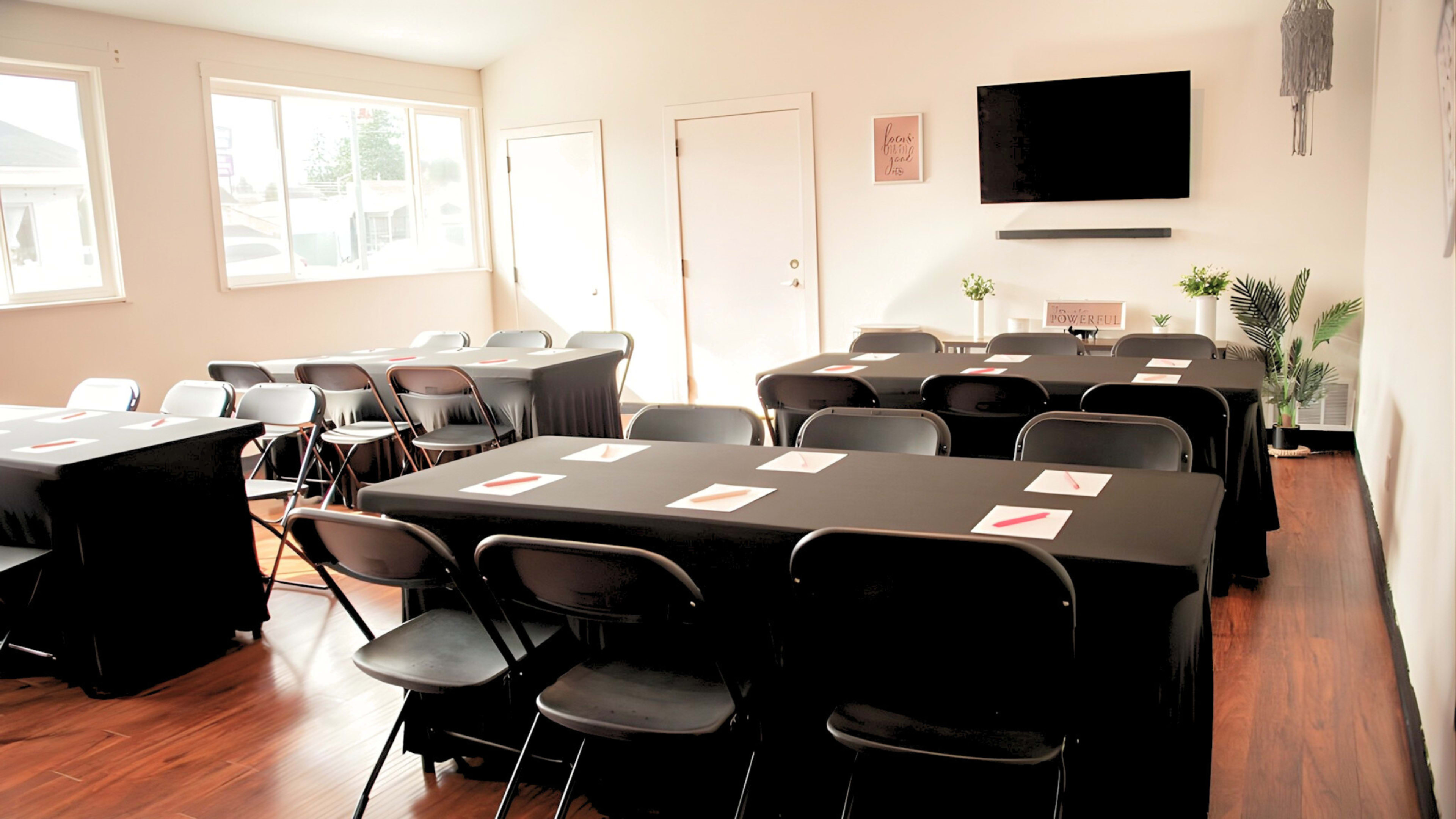 The image shows a well-lit meeting room arranged with several rows of black tables and chairs, with a television mounted on the wall and natural light streaming in through two windows.