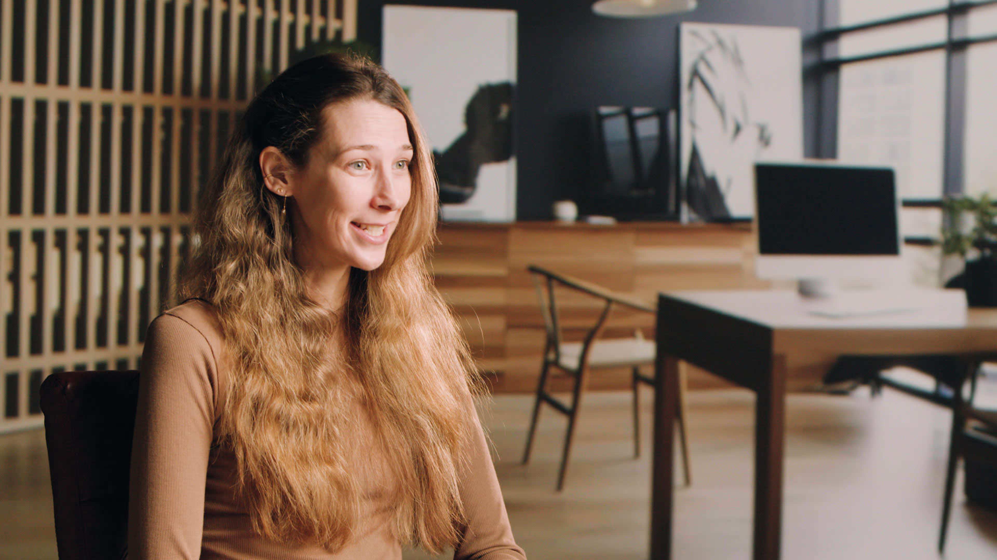 A woman with long, wavy hair sits in a modern office space with wooden furniture and large windows in the background.