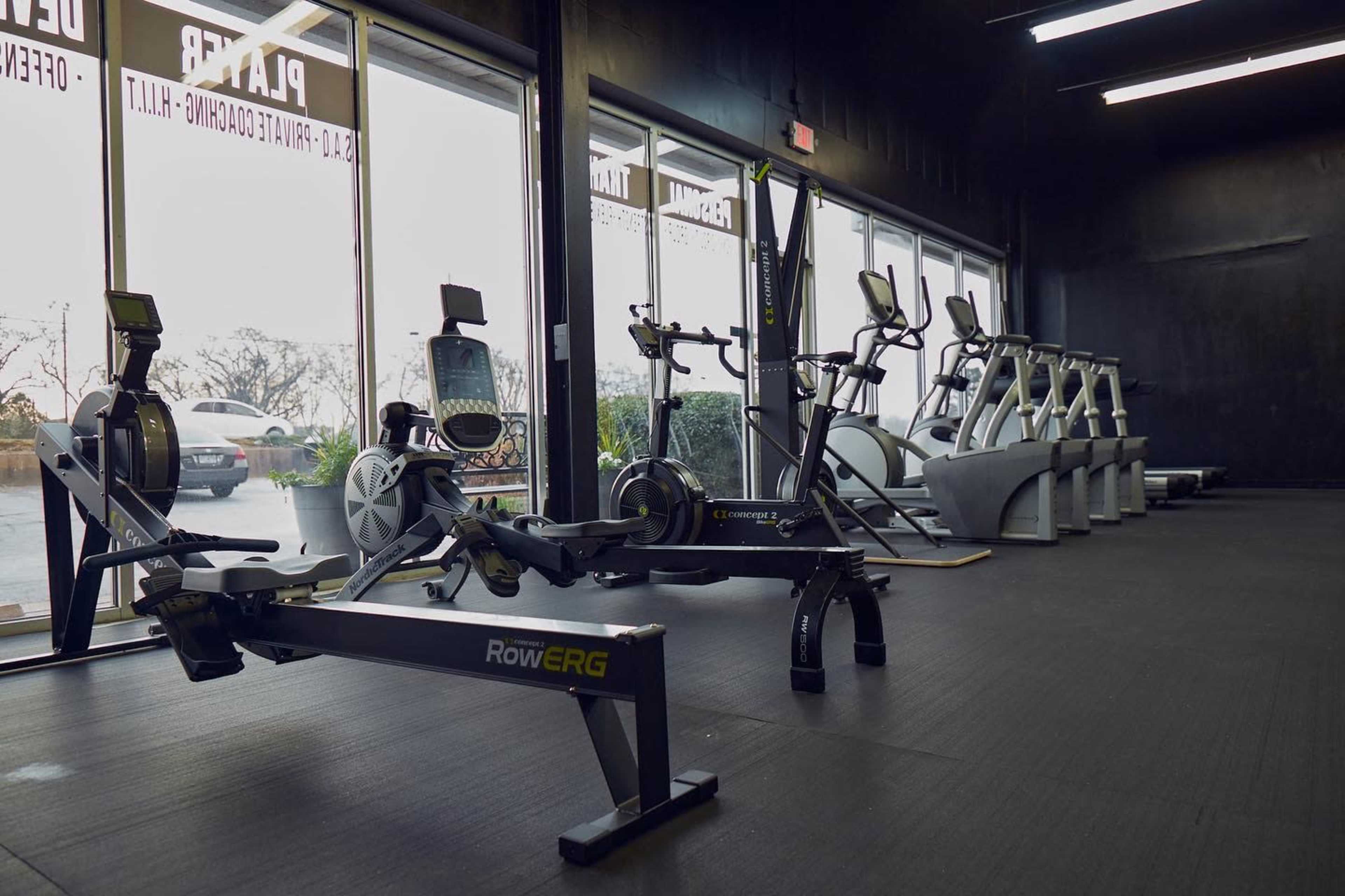 A fitness facility with rowing machines, stationary bikes, and elliptical machines arranged against large windows.