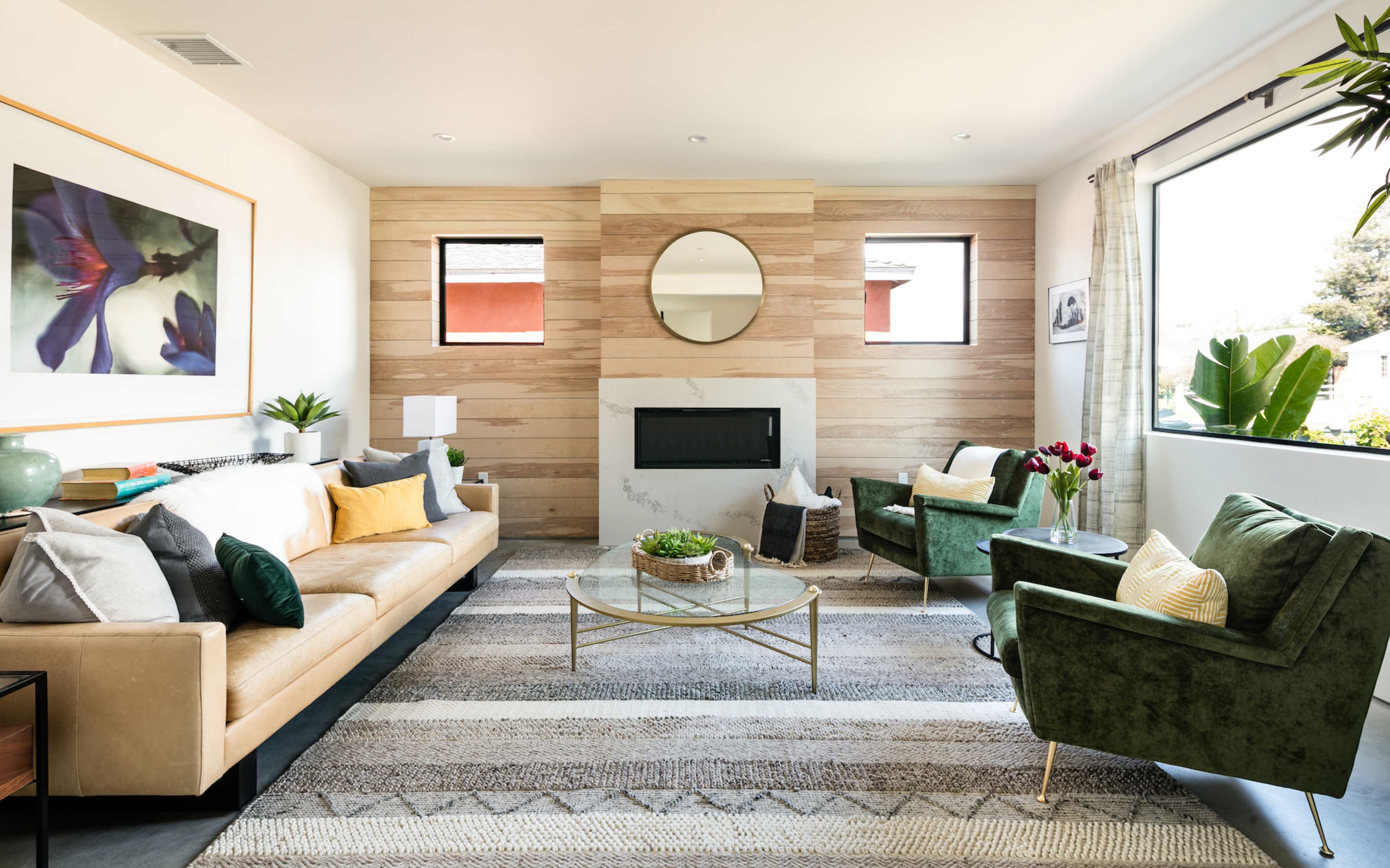 A modern living room features a beige sofa, green armchairs, a round mirror above a fireplace, and large windows letting in natural light.