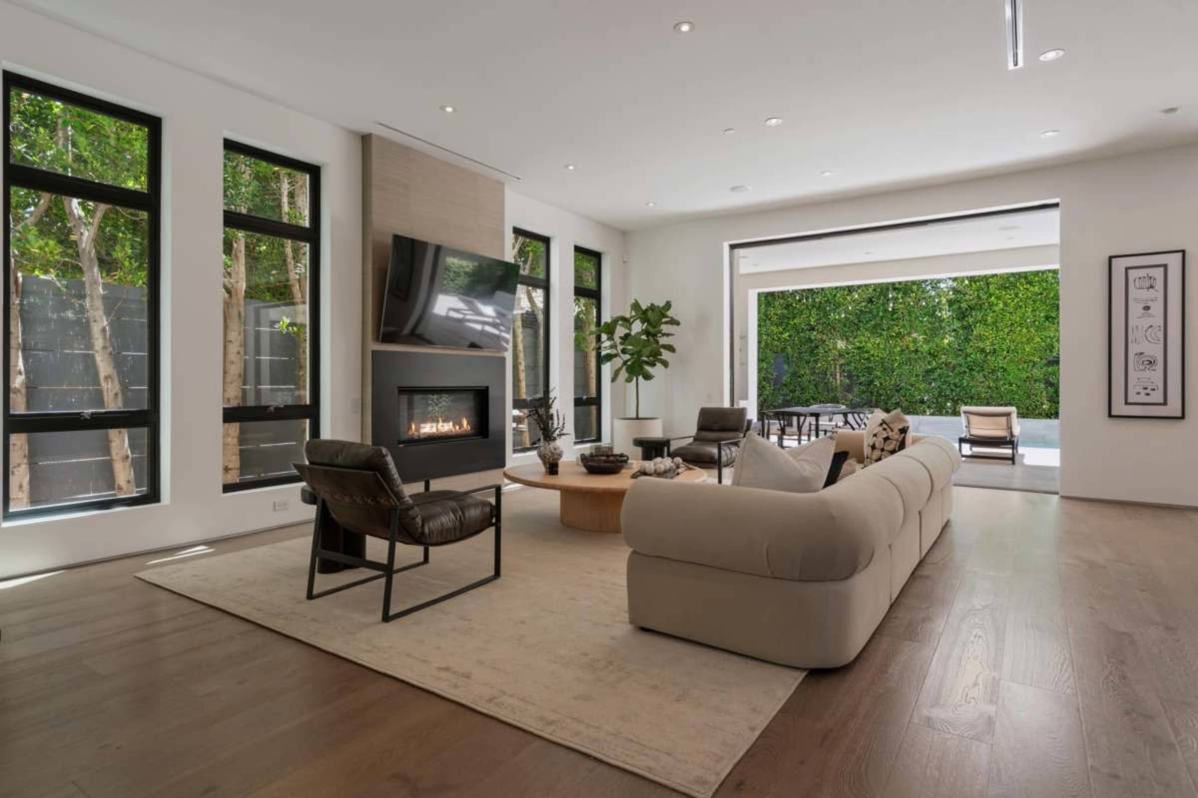 The image shows a modern living room with a large sofa, a coffee table, a fireplace, and floor-to-ceiling windows overlooking an outdoor patio area.