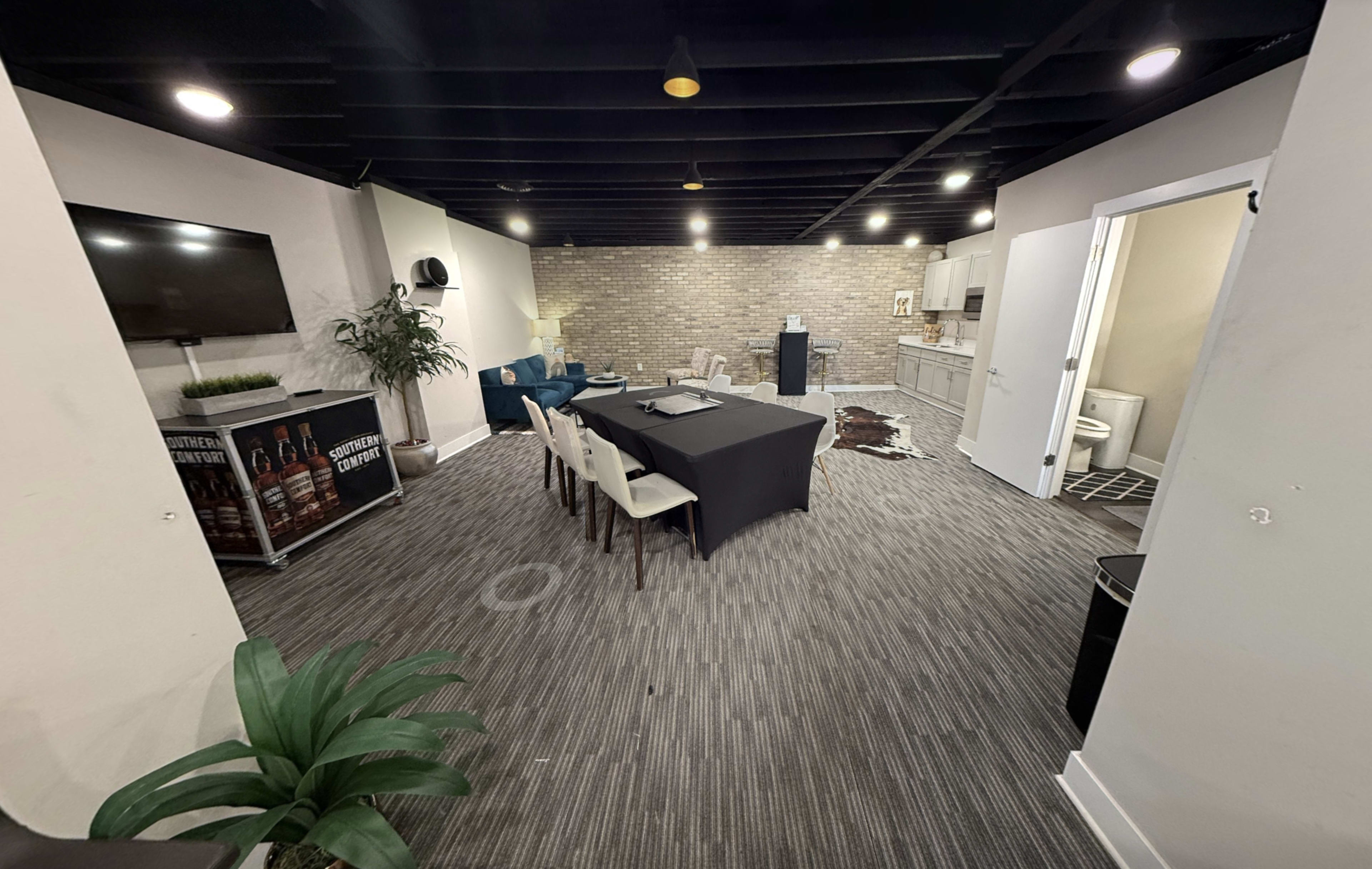 The image shows a modern conference room with a large table surrounded by chairs, and a kitchenette area in the background.