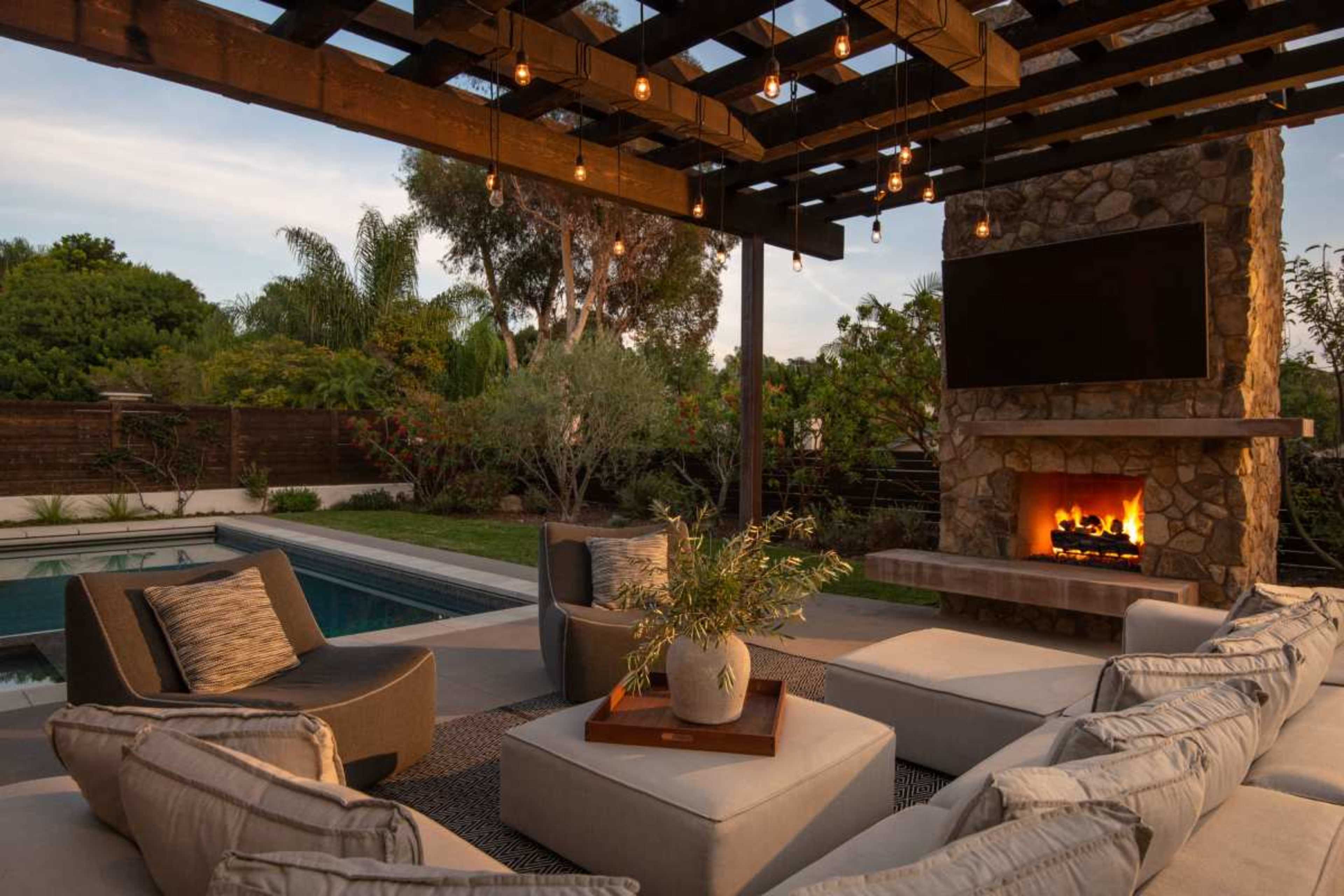 The image depicts a modern outdoor living space featuring a stone fireplace, a comfortable seating area, and a swimming pool surrounded by greenery.