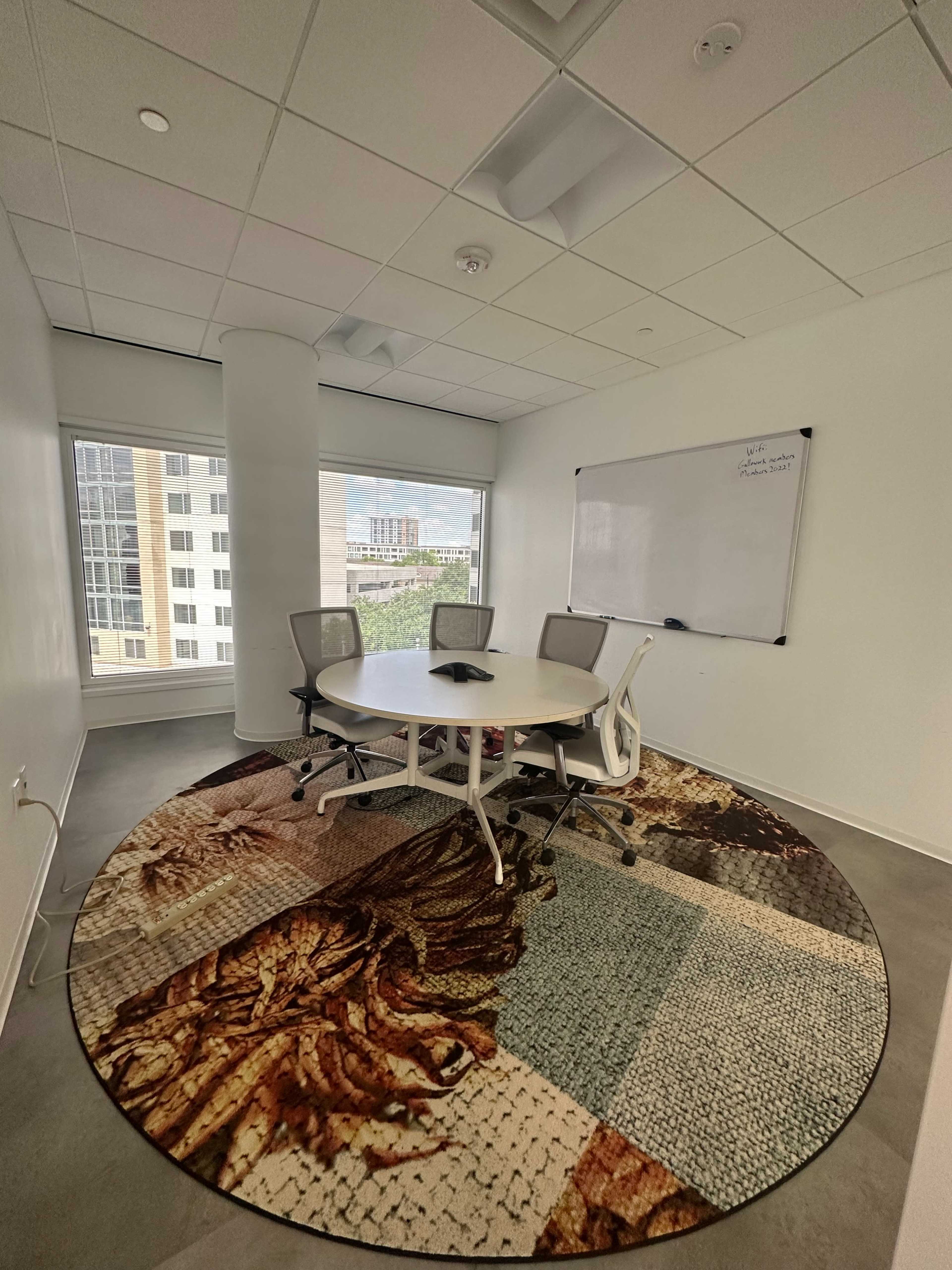 A conference room features a round rug with a detailed design, a whiteboard, a round table, and four chairs, with large windows providing natural light.