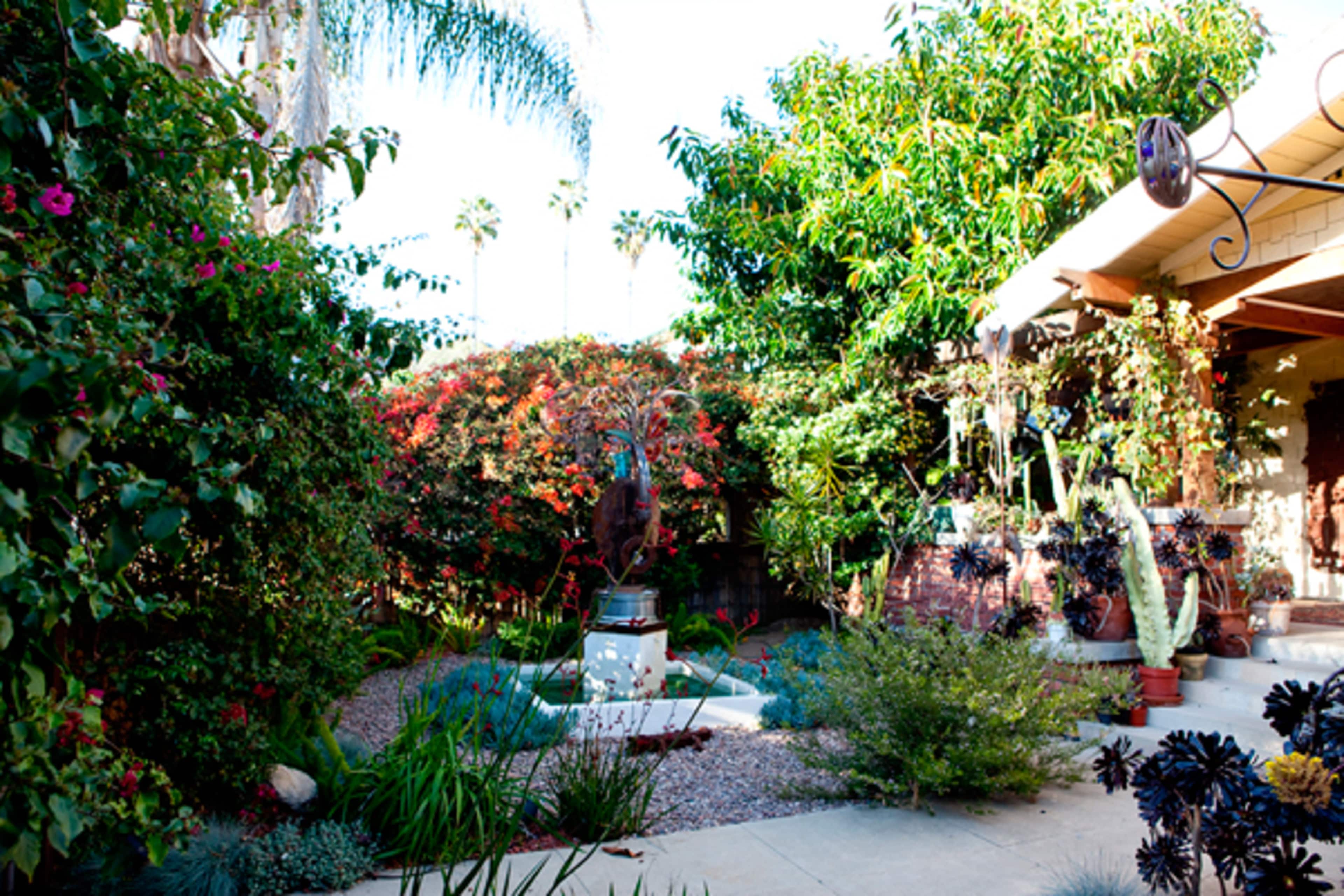 The image shows a garden with a variety of plants, a sculpture in the center, and a paved walkway surrounded by greenery.