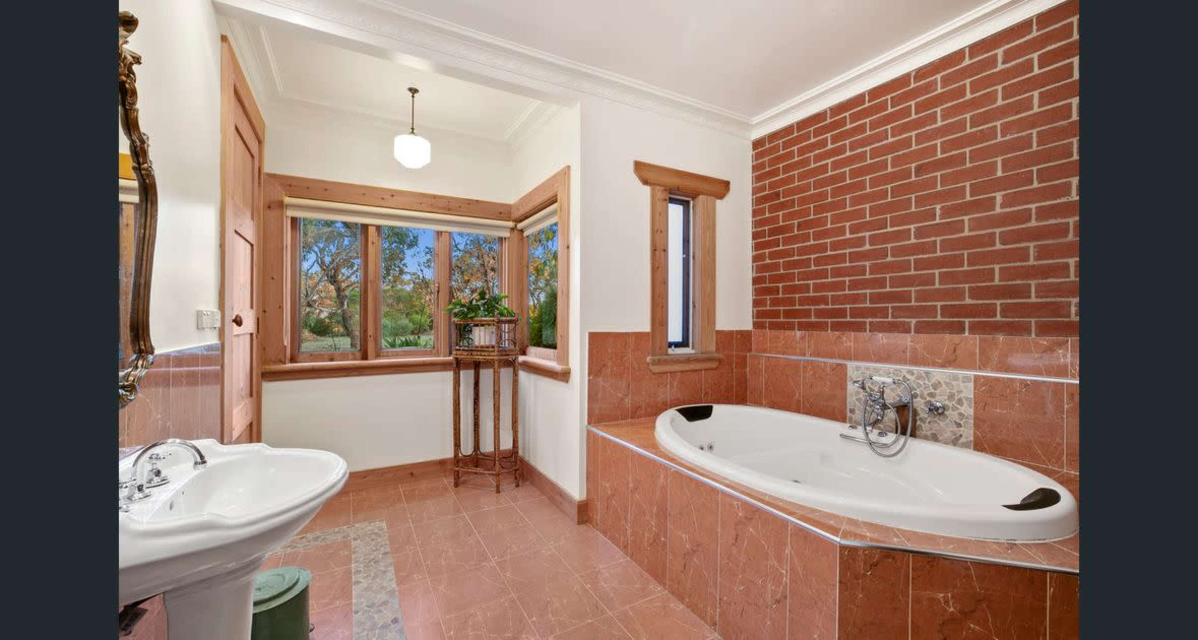 The image shows a spacious bathroom featuring a corner bathtub, a vintage sink, and a window with a view of greenery surrounded by brick and tiled walls.